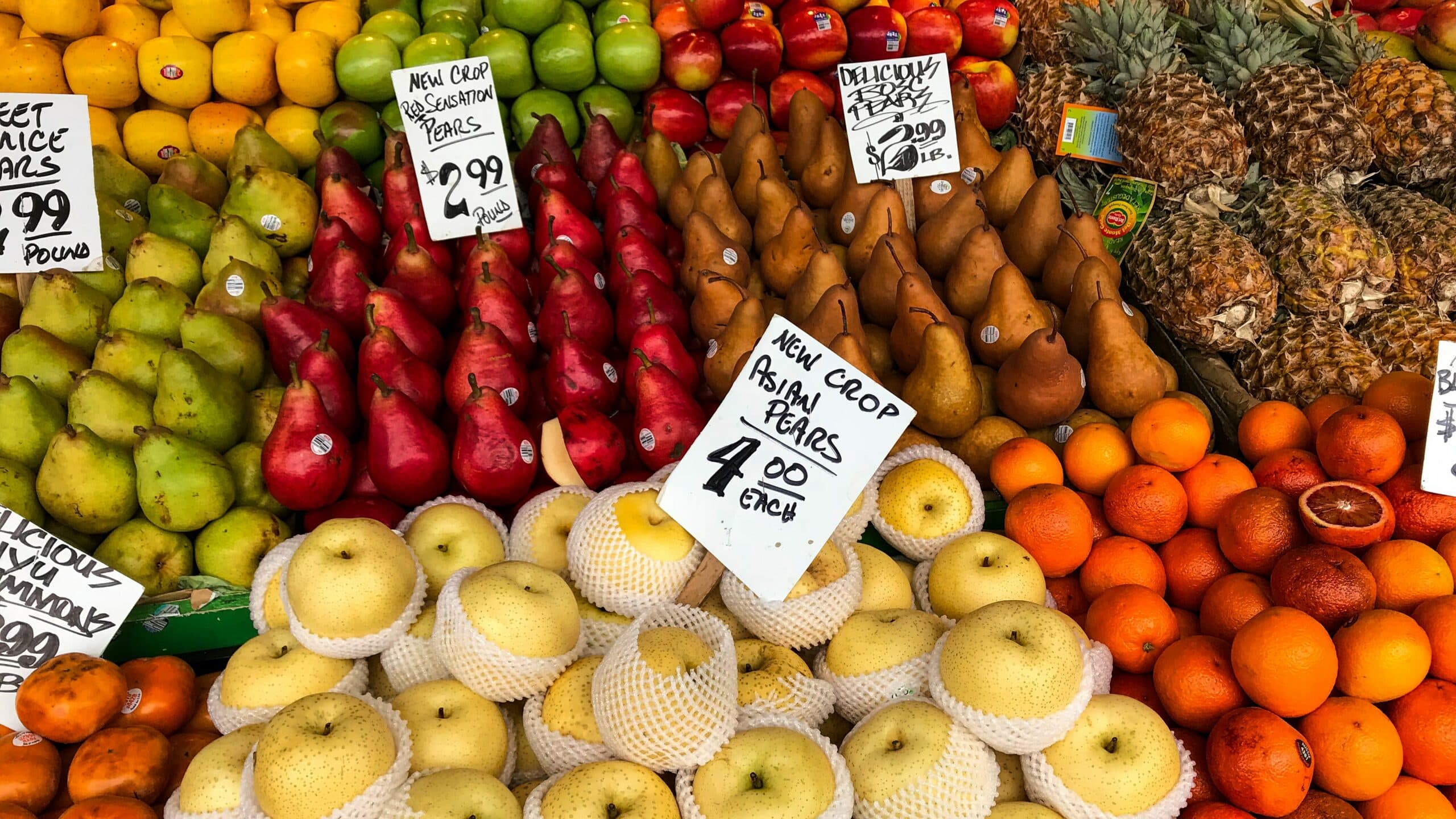 A colorful display of fruits at a market, featuring red and green pears, Asian pears wrapped in netting, pineapples, oranges, and apples. Handwritten price signs are visible above the fruit.
