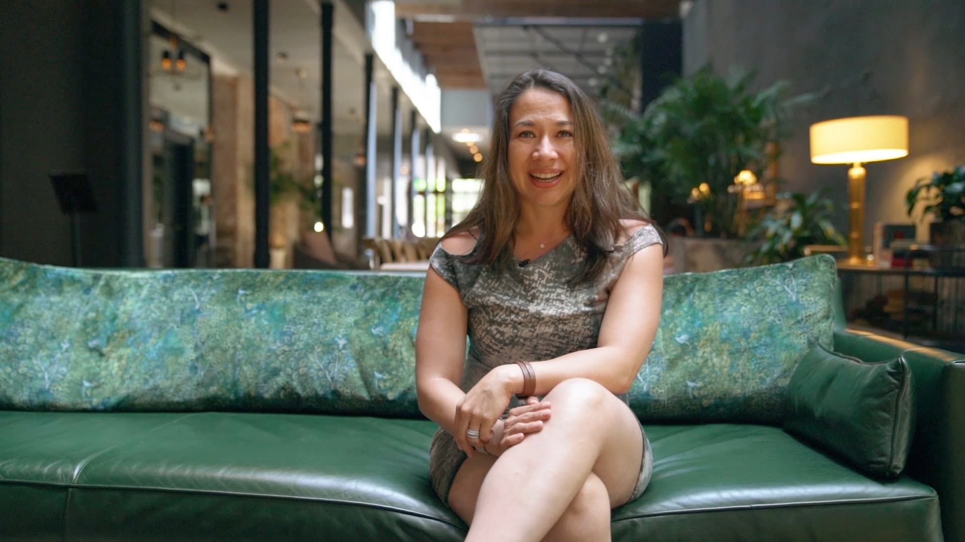 A woman with long brown hair sits on a green sofa in a modern, stylish interior with plants and soft lighting. She is smiling and wearing a patterned dress, with her legs crossed.