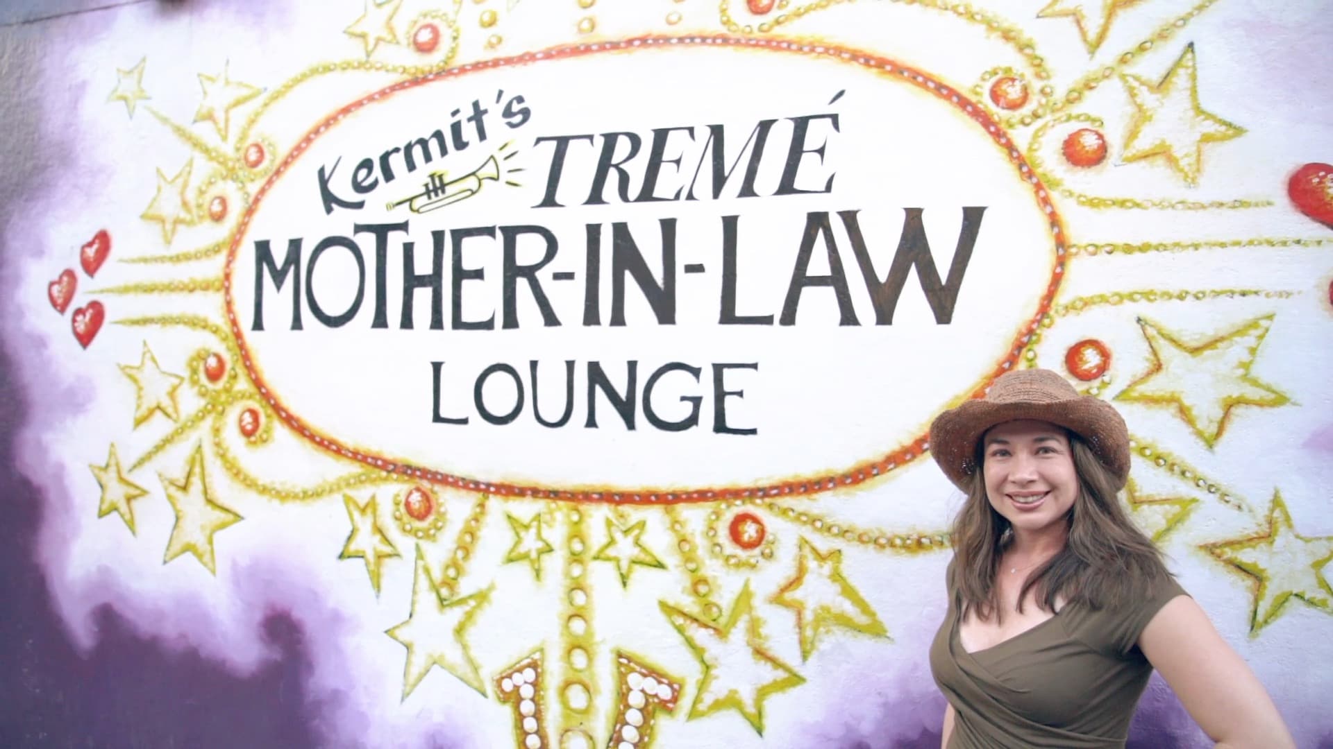 A woman wearing a brown hat and olive dress stands smiling in front of a colorful mural that reads Kermits Tremé Mother-In-Law Lounge. The mural is decorated with stars and musical motifs on a purple background.