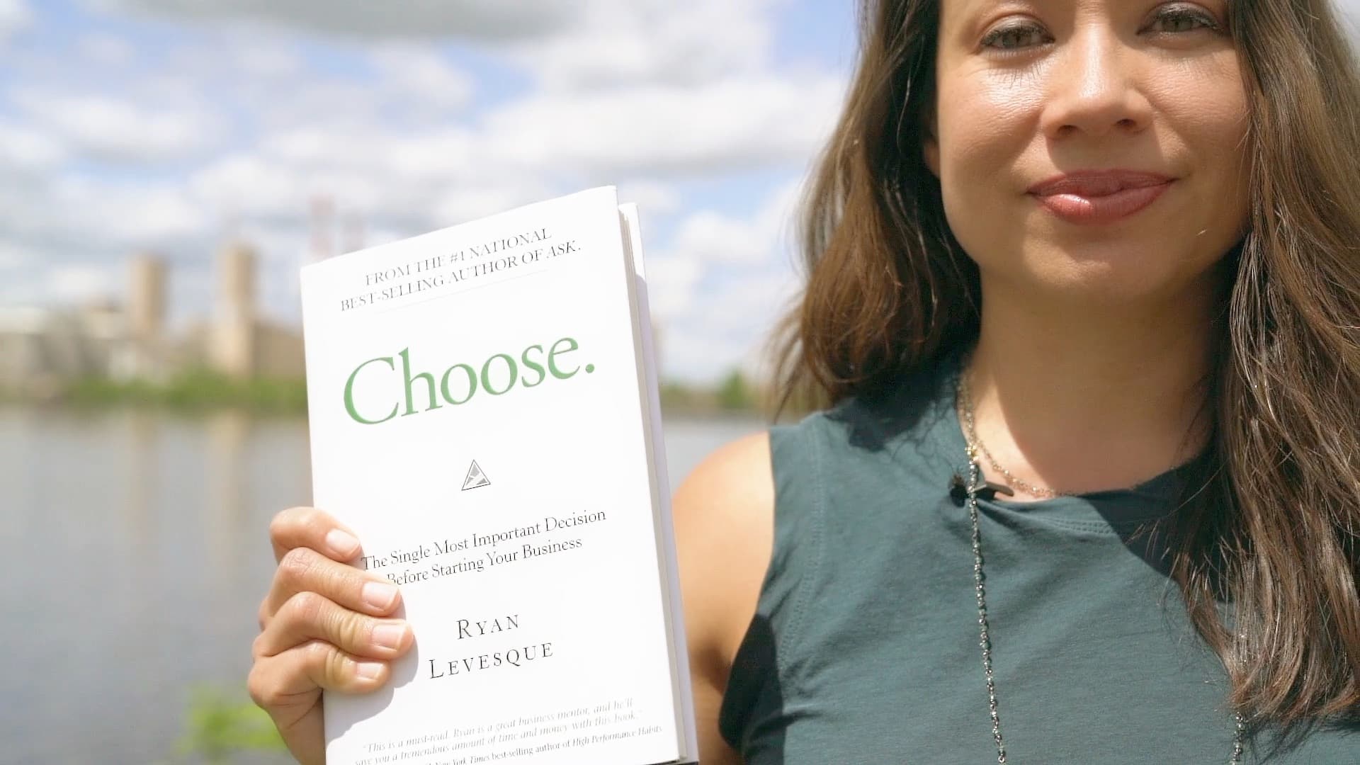 A woman outdoors holds up the book Choose: The Single Most Important Decision Before Starting Your Business by Ryan Levesque. Trees and a body of water are visible in the background under a cloudy sky.