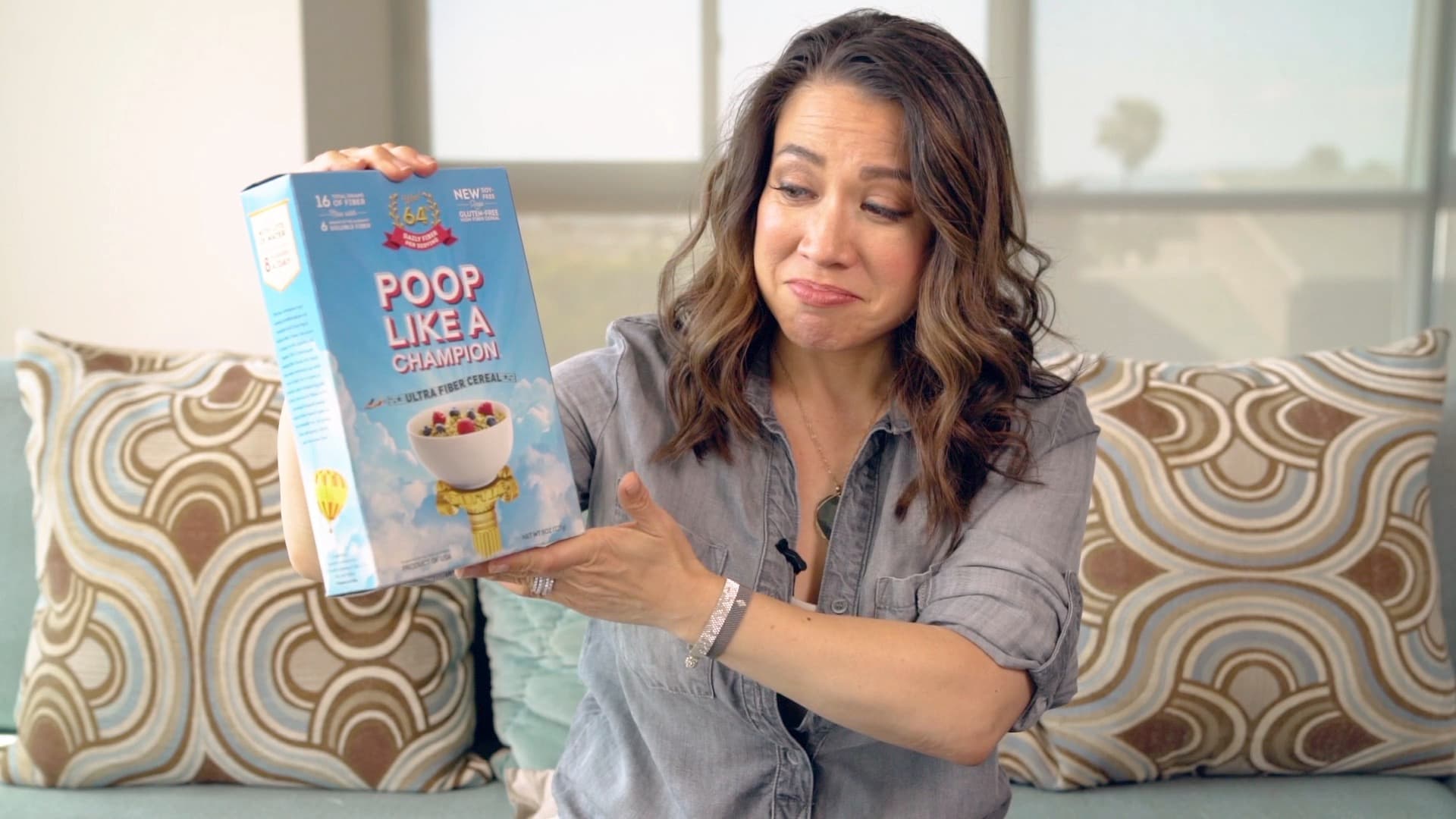 A woman sits on a couch, holding a box labeled Poop Like A Champion. She appears amused and slightly skeptical. The background includes patterned pillows and a window.