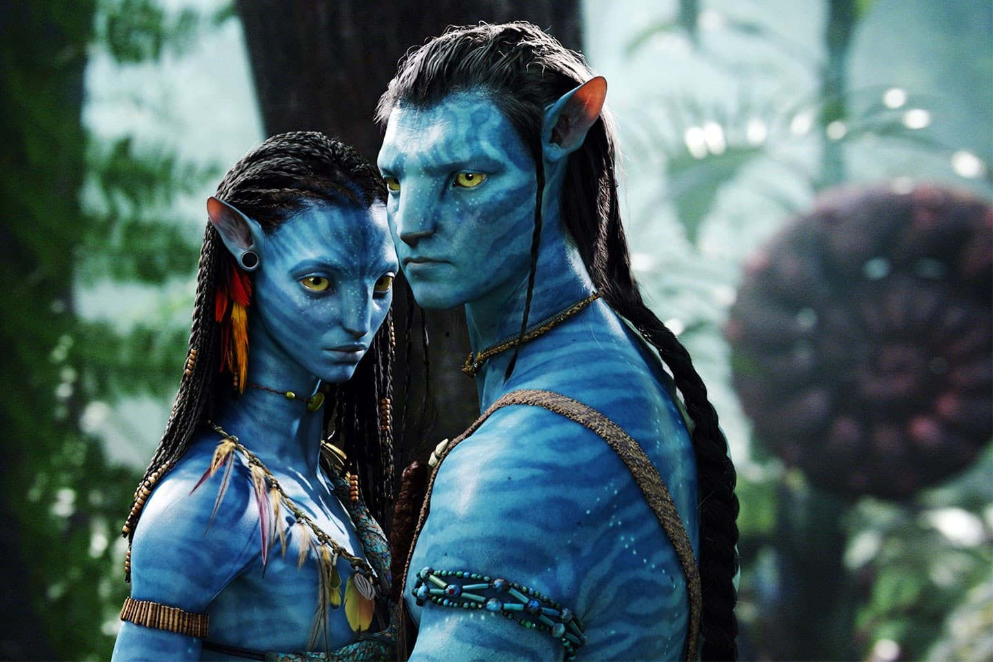 Two blue-skinned, humanoid figures with pointed ears and braided hair stand together in a lush, green forest environment. The figure on the left has beaded jewelry and colorful feathers, while the figure on the right wears simple adornments.