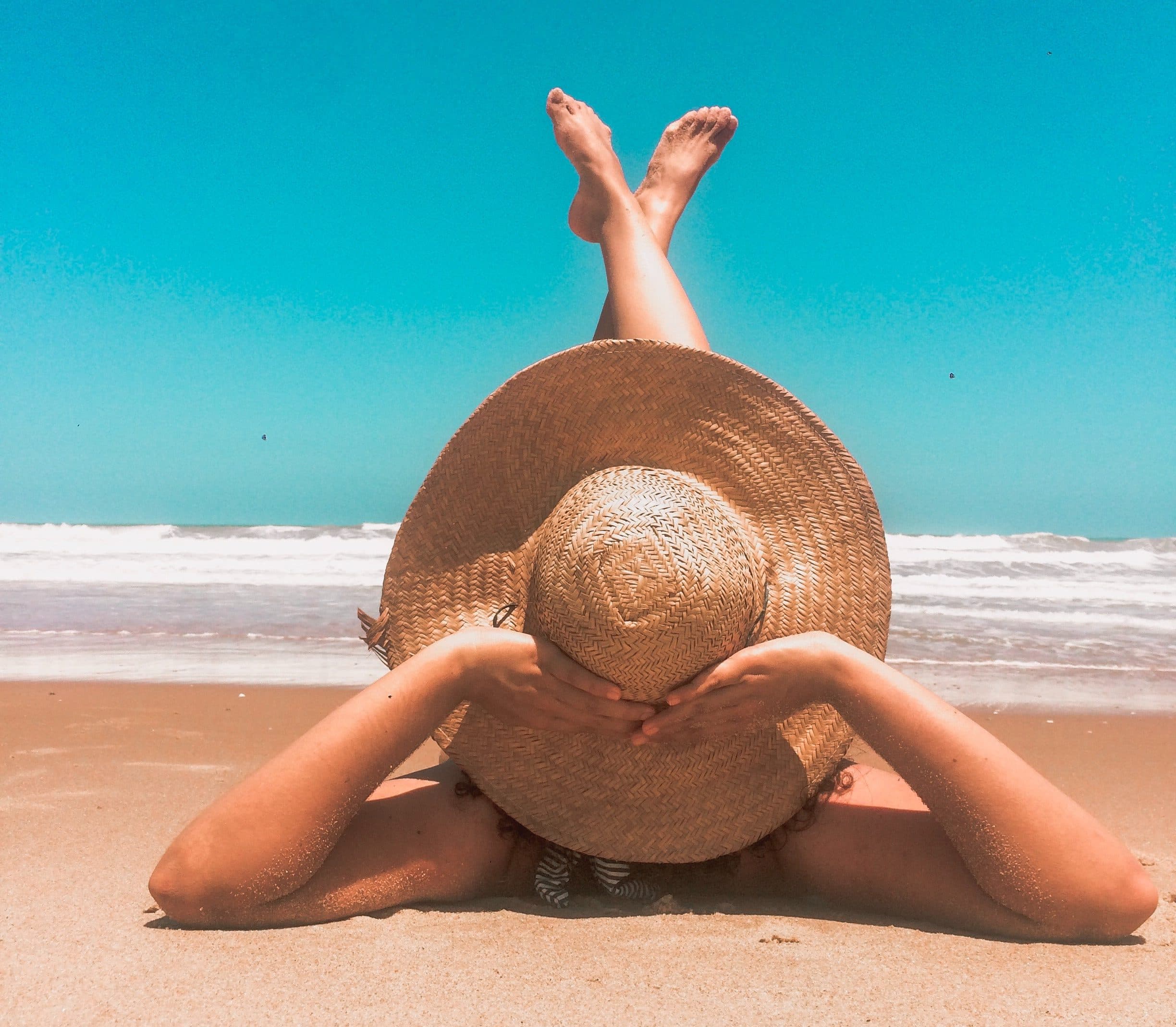 A person lies on a sandy beach, face down, with legs raised in the air. They wear a large straw hat and have their hands resting on the hats brim. The ocean and a clear blue sky are in the background.
