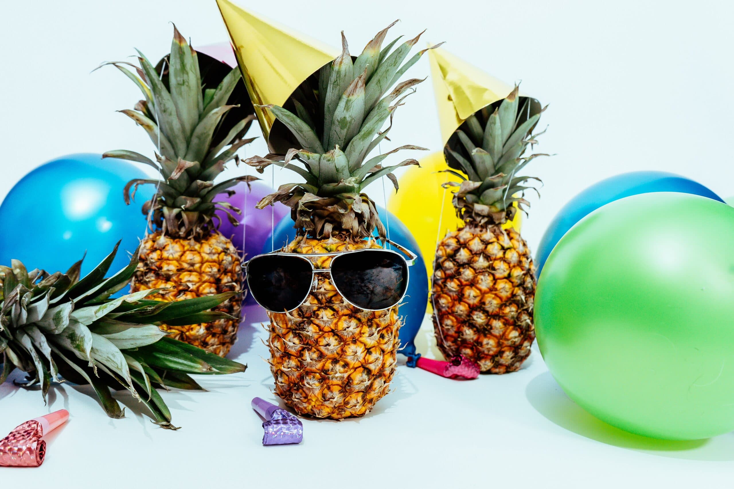 Three pineapples with party hats and sunglasses are surrounded by colorful balloons and party blowers. The playful scene suggests a festive, tropical party atmosphere.