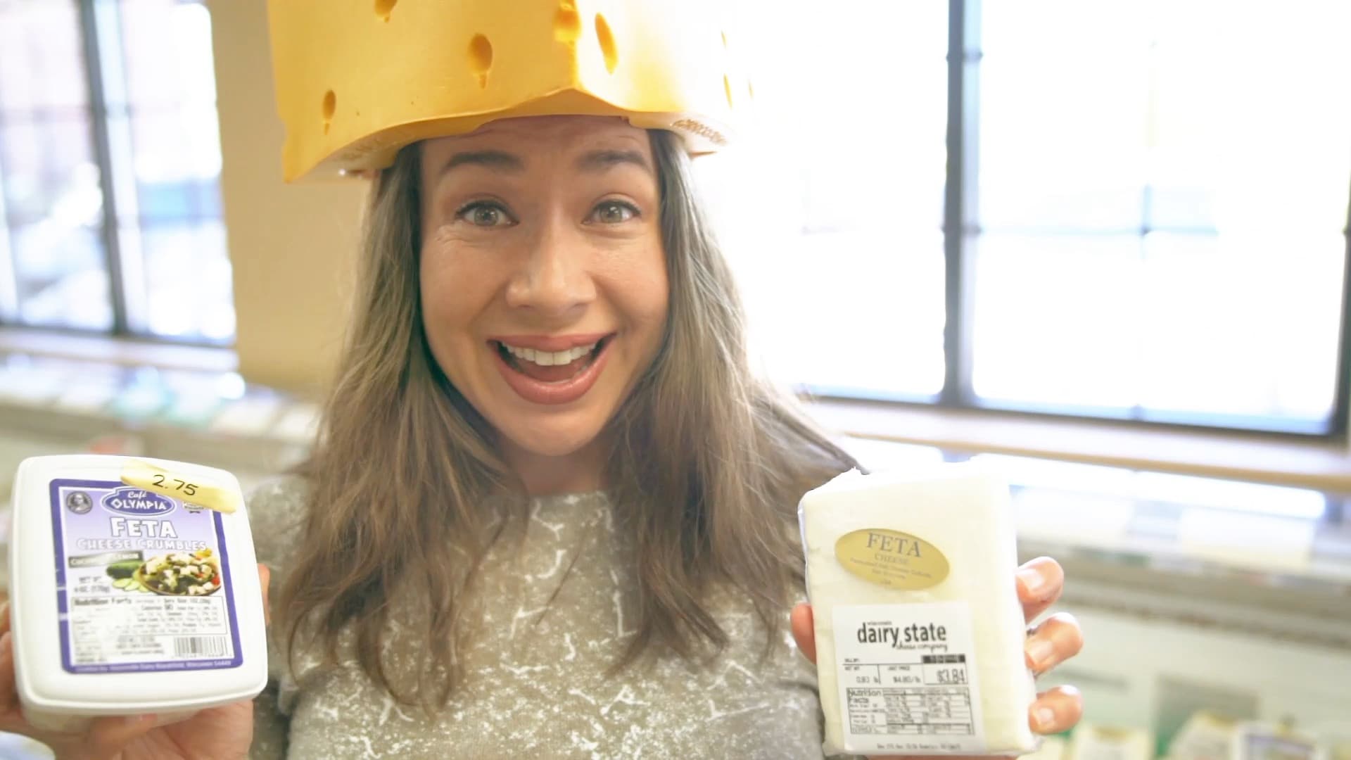 A person with long hair is smiling and wearing a cheese-shaped hat. They are holding containers of feta cheese in both hands, standing indoors in front of a window.