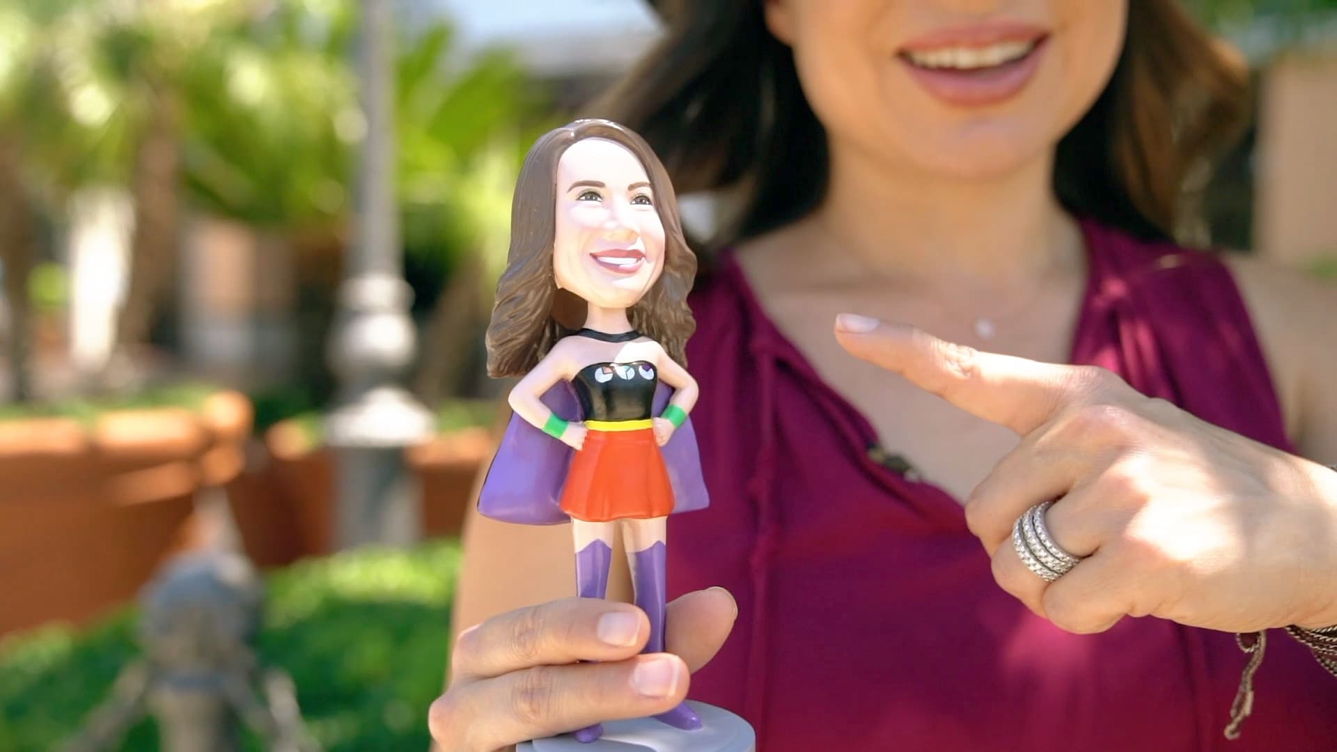 A person holds a superhero-themed bobblehead with a cape and skirt, smiling broadly while pointing at it. The background features greenery and a blurred outdoor setting.