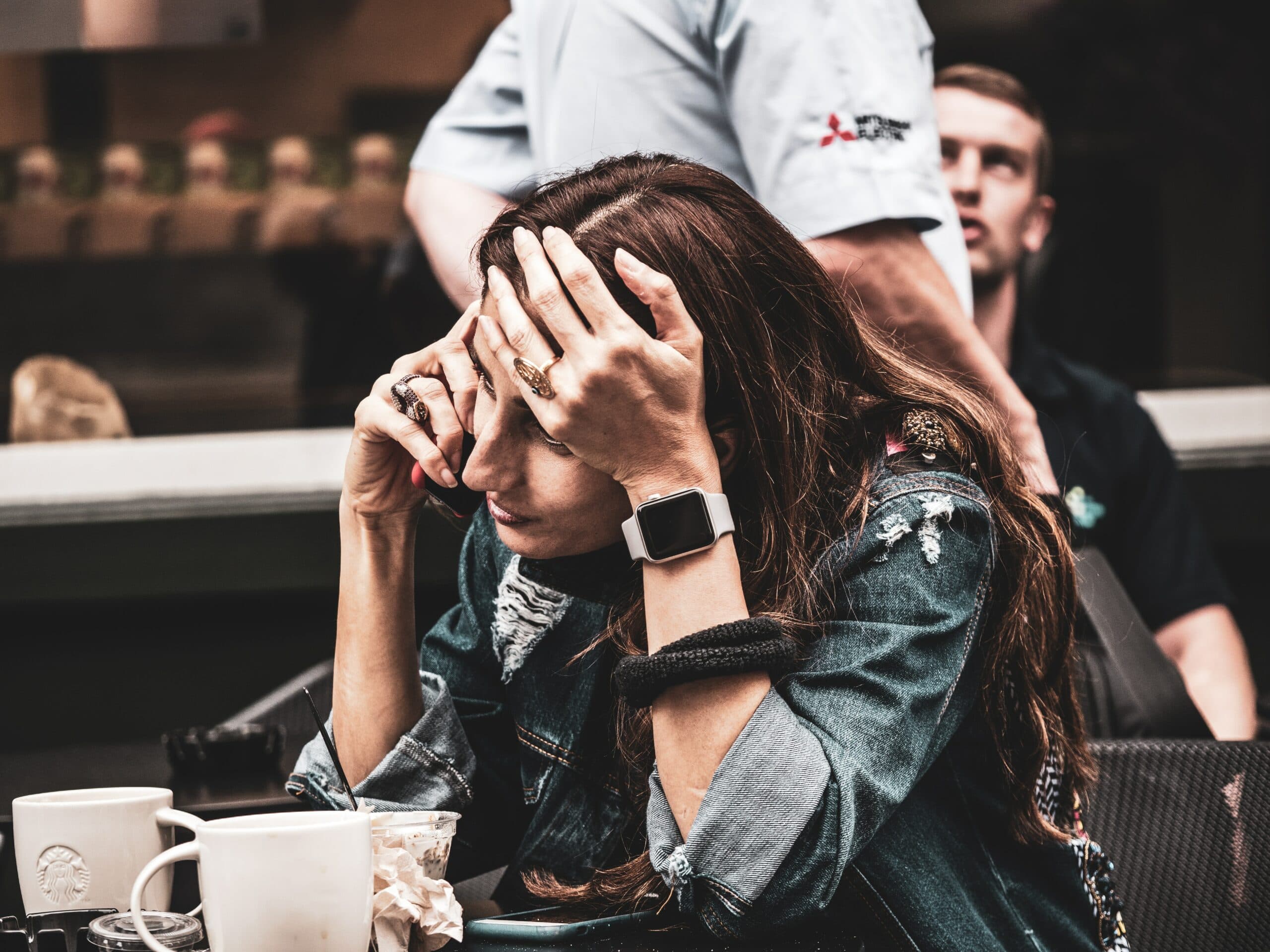 A woman sits at a table with her hands on her forehead, appearing stressed. She wears a denim jacket and has a smartwatch on her wrist. There are several cups and a crumpled napkin on the table, and a person stands behind her.