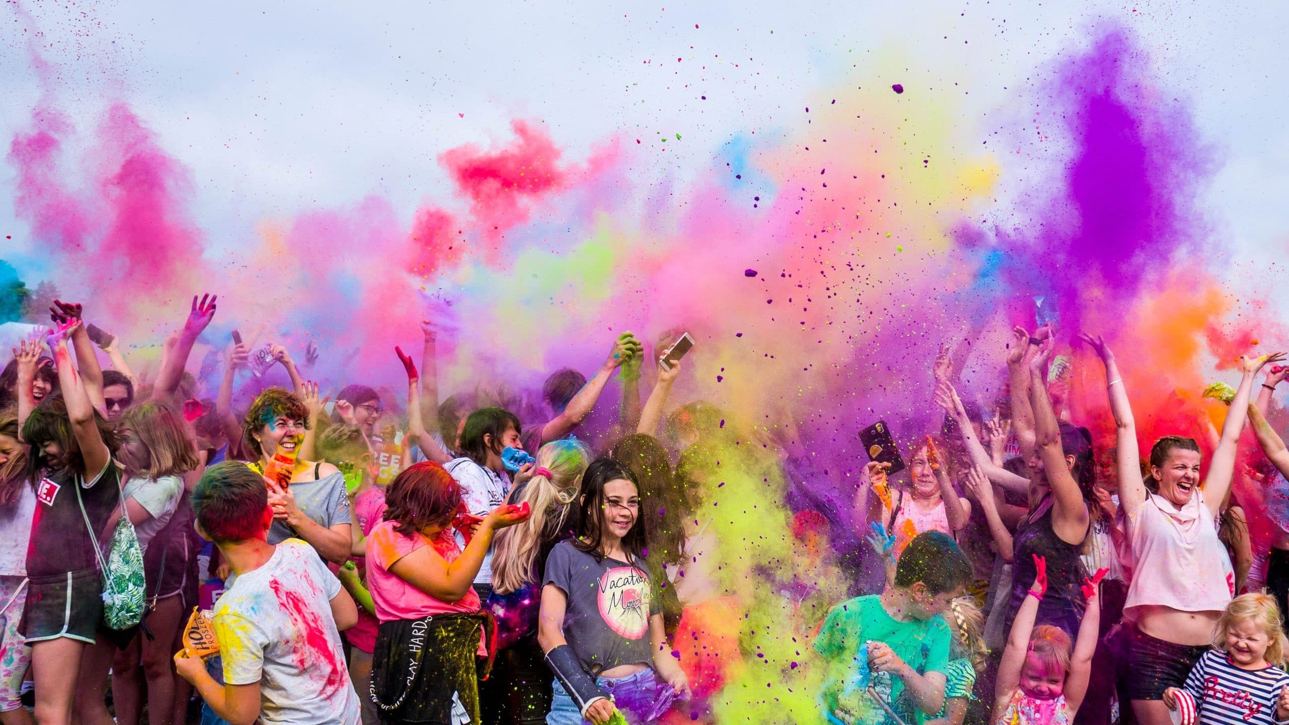 A joyful crowd is celebrating by throwing vibrant colored powders into the air at an outdoor festival. The sky is filled with a mix of pink, purple, yellow, and green hues. People are smiling and taking pictures amidst the lively atmosphere.
