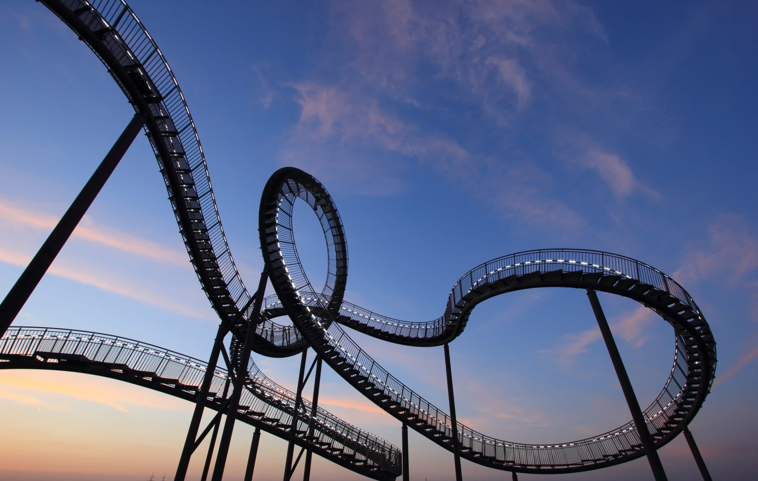 A looping roller coaster structure silhouetted against a twilight sky with pink and blue hues. The intricate design includes loops and twists, elevated on tall supports, creating a dramatic contrast against the evening backdrop.