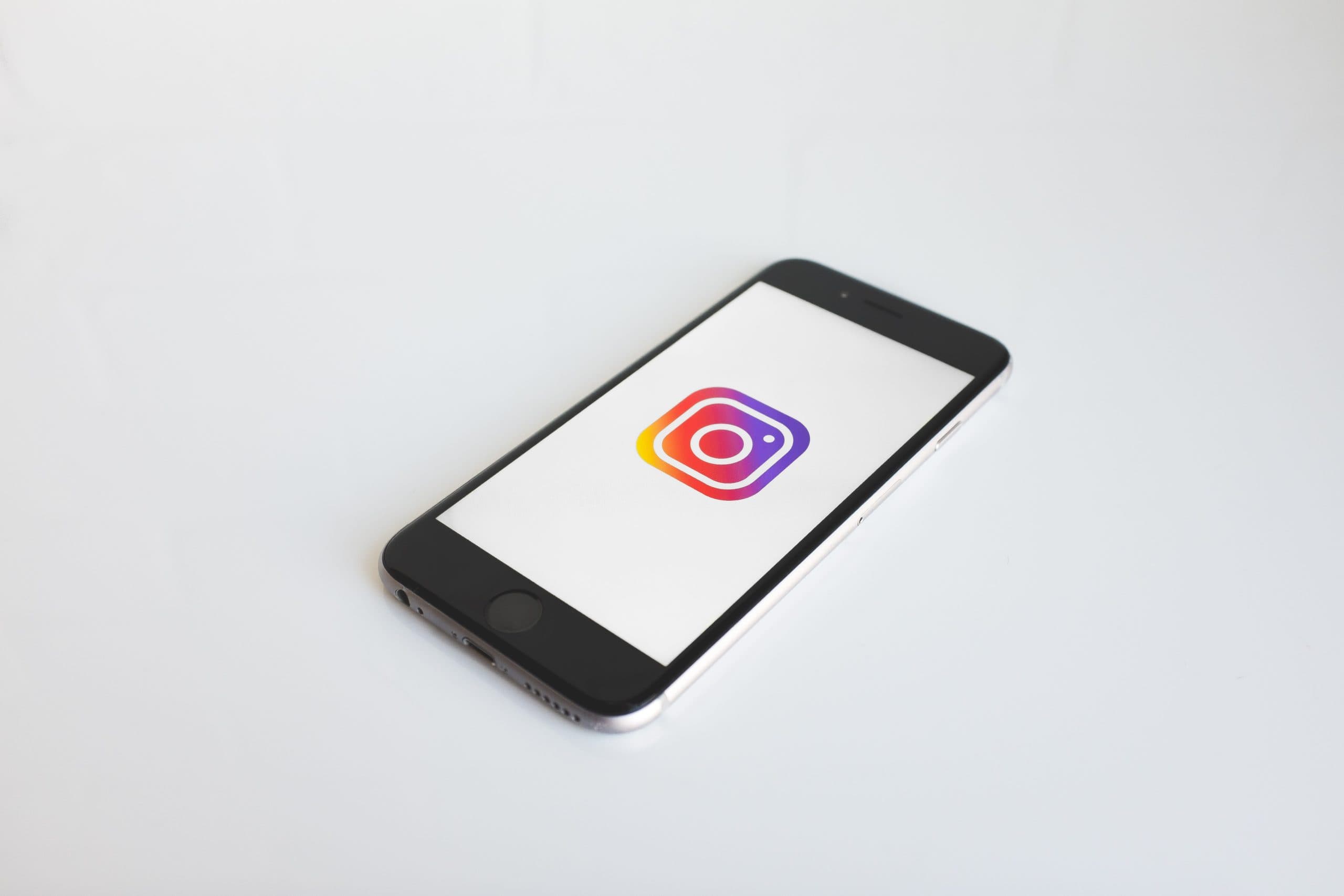 A smartphone displaying the Instagram logo on its screen, placed on a white surface. The phone is positioned at a slight angle, with the logo clearly visible in vibrant colors.