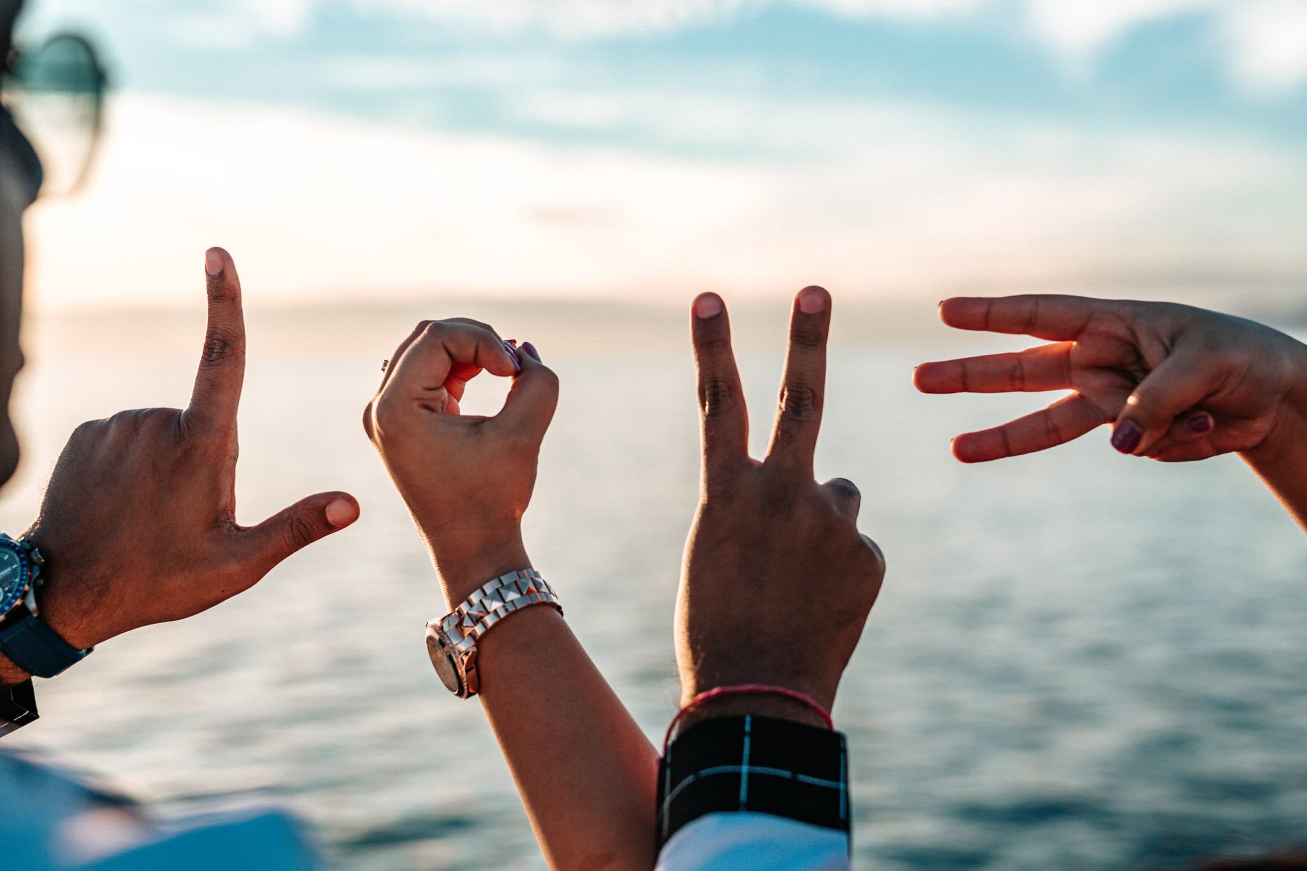 Four hands spelling LOVE in American Sign Language against a backdrop of a calm sea and a bright sky. The hands form the letters L, O, V, and E, from left to right, with sunlight reflecting on the water.