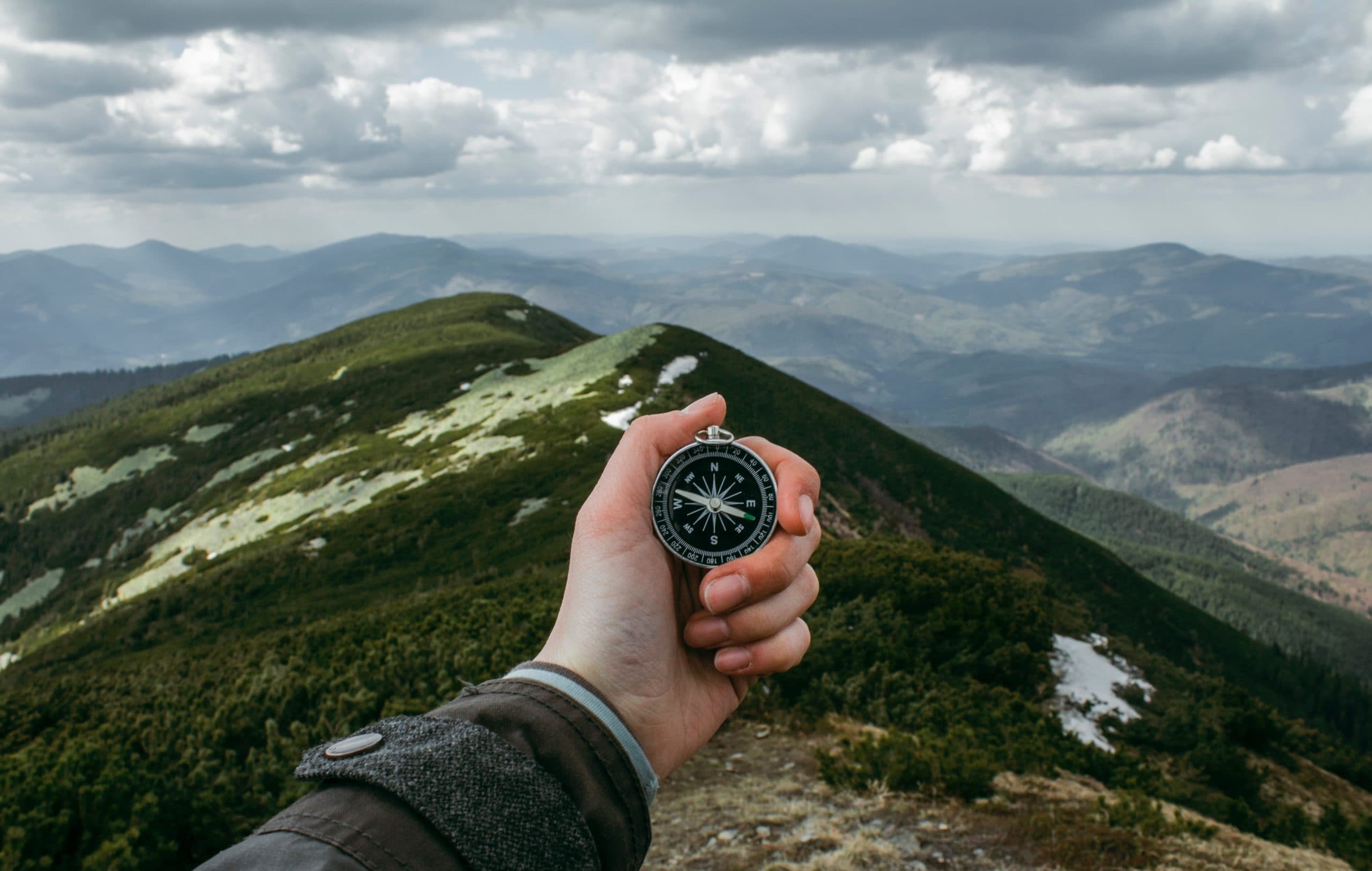 A hand holding a compass against a backdrop of a mountainous landscape. The sky is cloudy, and the rolling green hills stretch into the distance, creating a sense of adventure and exploration.