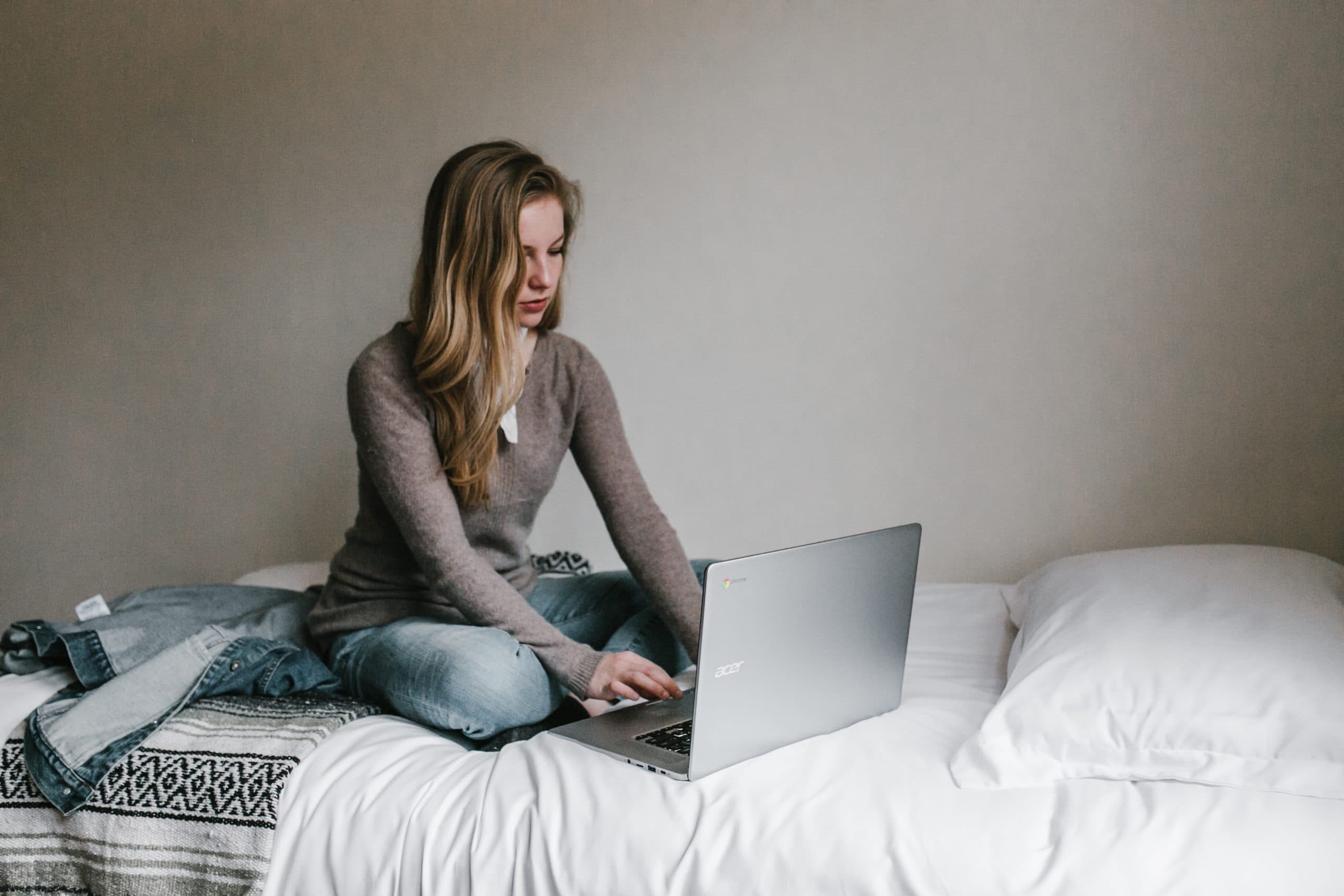 A person sitting on a bed, using a laptop. They wear a brown sweater and jeans, and theres a jacket and blanket beside them. The background is a plain wall and pillows are visible on the bed.