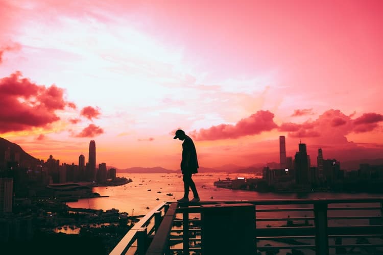 A person in silhouette stands on a rooftop railing against a vibrant red and pink sky. The city skyline and a harbor with boats are visible below, creating a dramatic urban sunset scene.