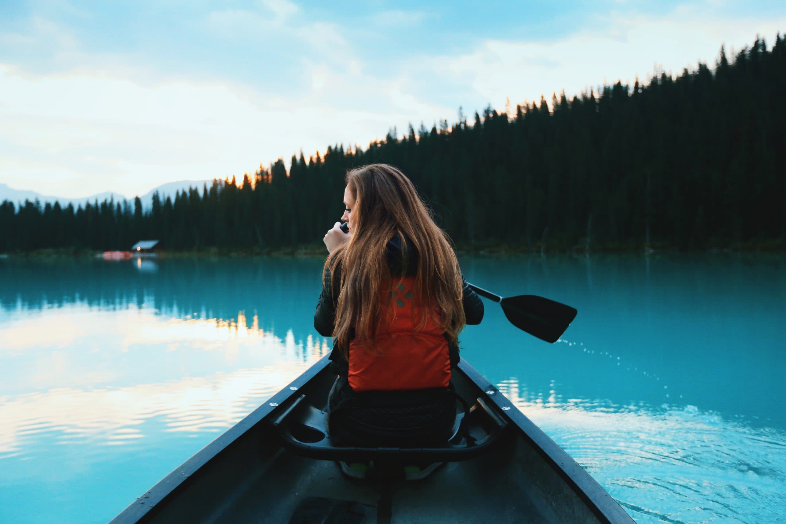 A person with long hair wearing a red life vest is sitting in a canoe, paddling on a serene, turquoise lake surrounded by a dense forest. The sky is lightly clouded, and the calm water reflects the natural scenery.