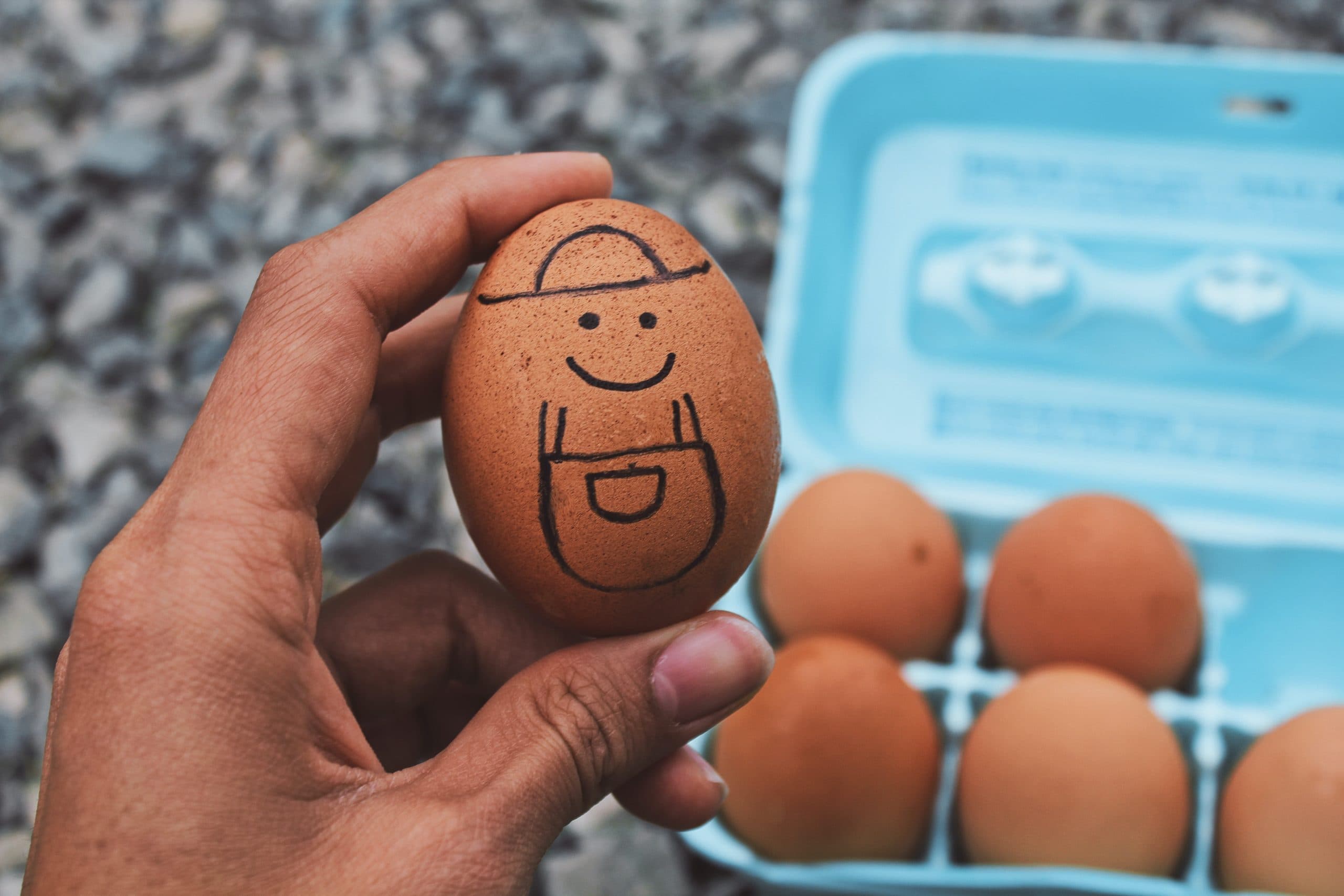 A hand holding a brown egg with a smiley face and a cap drawn on it, resembling a character wearing overalls. In the background, more eggs are visible in a blue carton on a gravel surface.