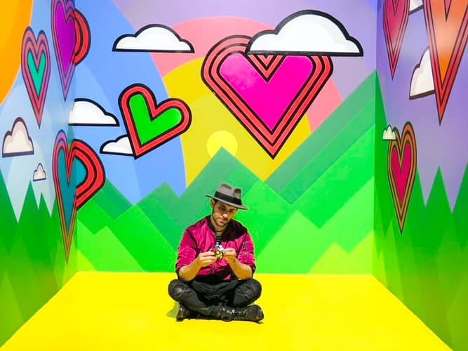 Person sitting on a bright yellow floor in a colorful room with heart and cloud designs on the walls. The person wears a hat and is holding a Rubiks cube. The background features green mountains and a sun.