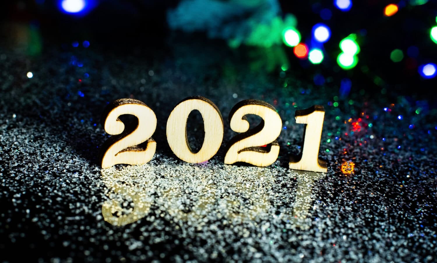 Wooden 2021 figures stand upright on a glittery surface, surrounded by colorful, blurred lights in the background, creating a festive atmosphere.