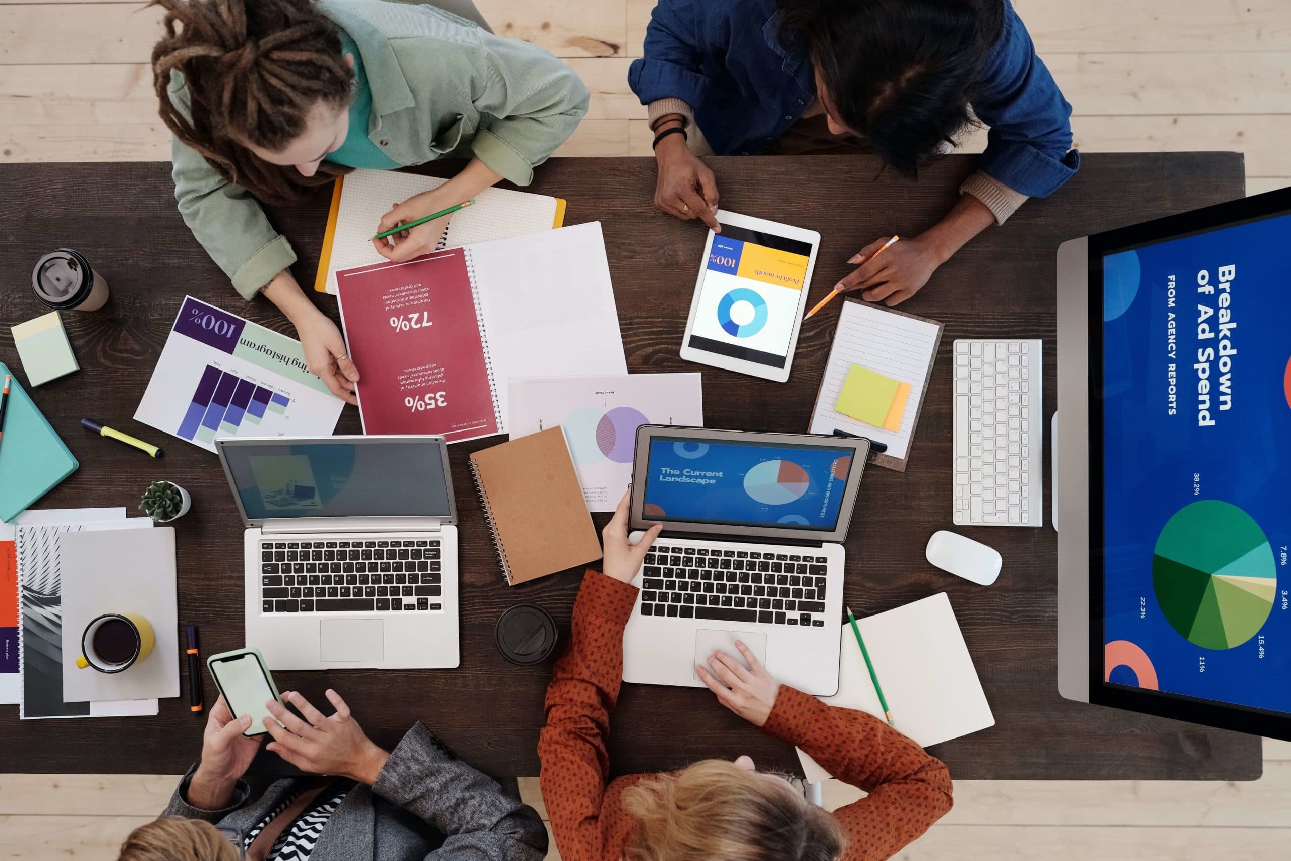 Aerial view of a group of people collaborating at a table with laptops, tablets, notebooks, and charts. The screen displays graphs and the text Breakdown of Ad Spend. The setting is an office or workspace.