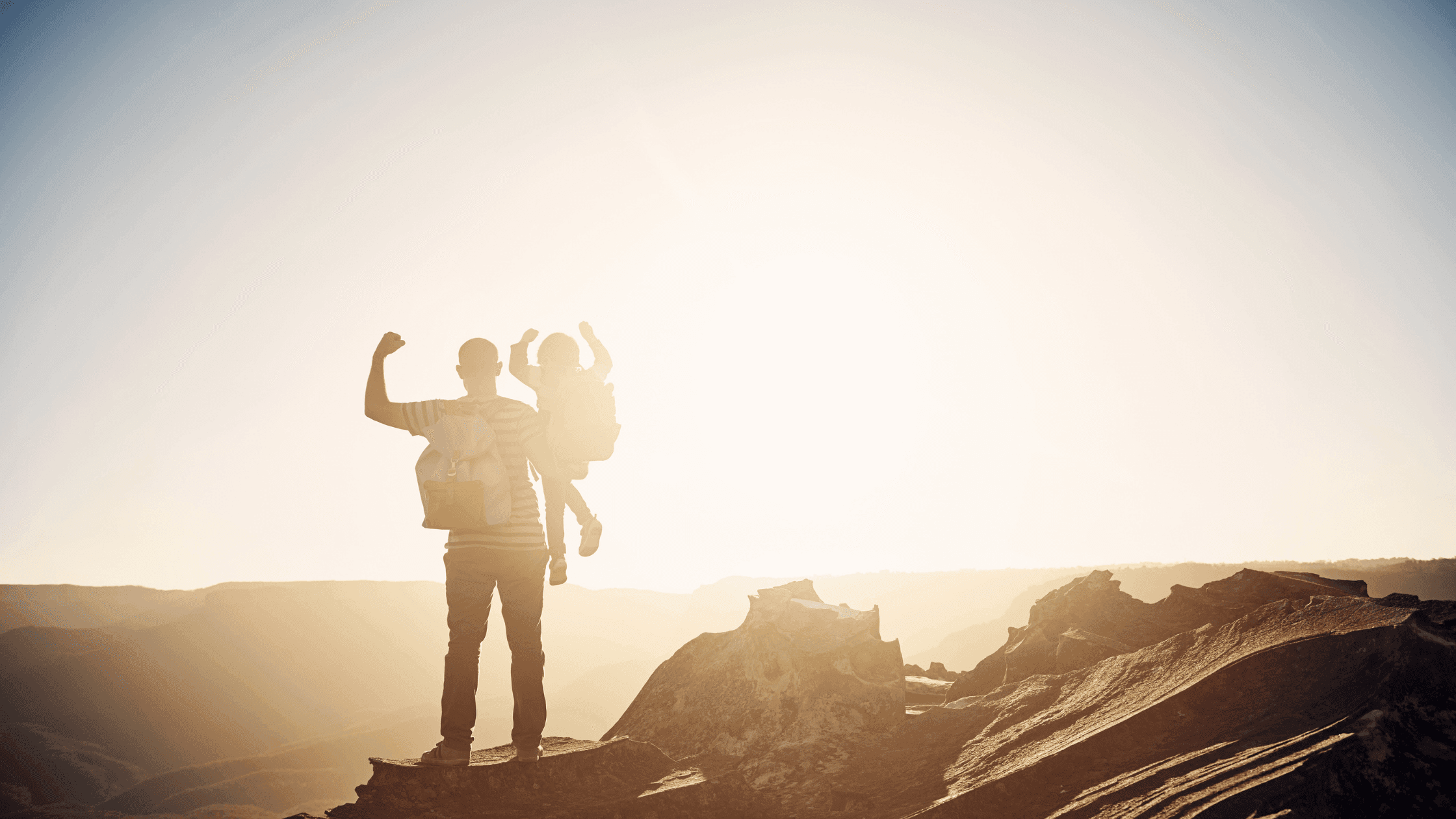 A person stands on rocky terrain at sunrise, holding a child on their shoulders. The silhouettes are set against a bright sky, symbolizing adventure and togetherness.