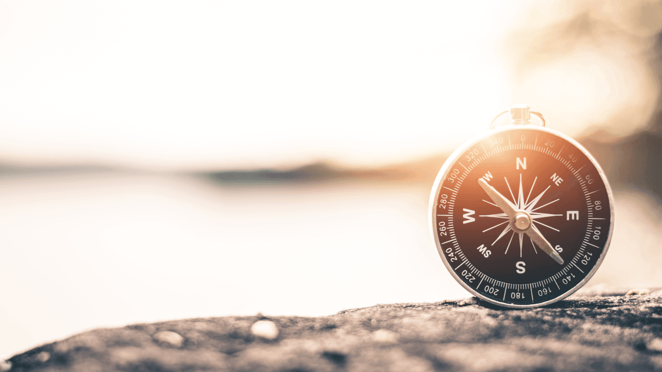 A compass rests on a rock with a blurred, sunlit background. The compass needle points toward the cardinal directions, capturing a serene and adventurous atmosphere.