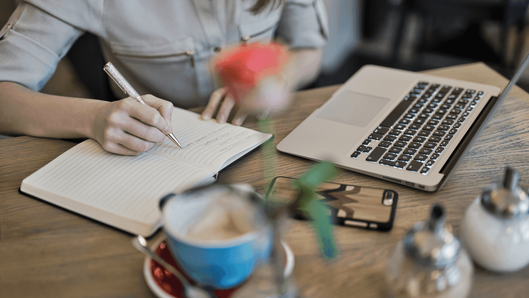 A person writes in a notebook with a pen next to an open laptop on a wooden table. A blurred rose, a smartphone, and a mug with a saucer are also visible. The setting appears to be a cafe or a casual workspace.