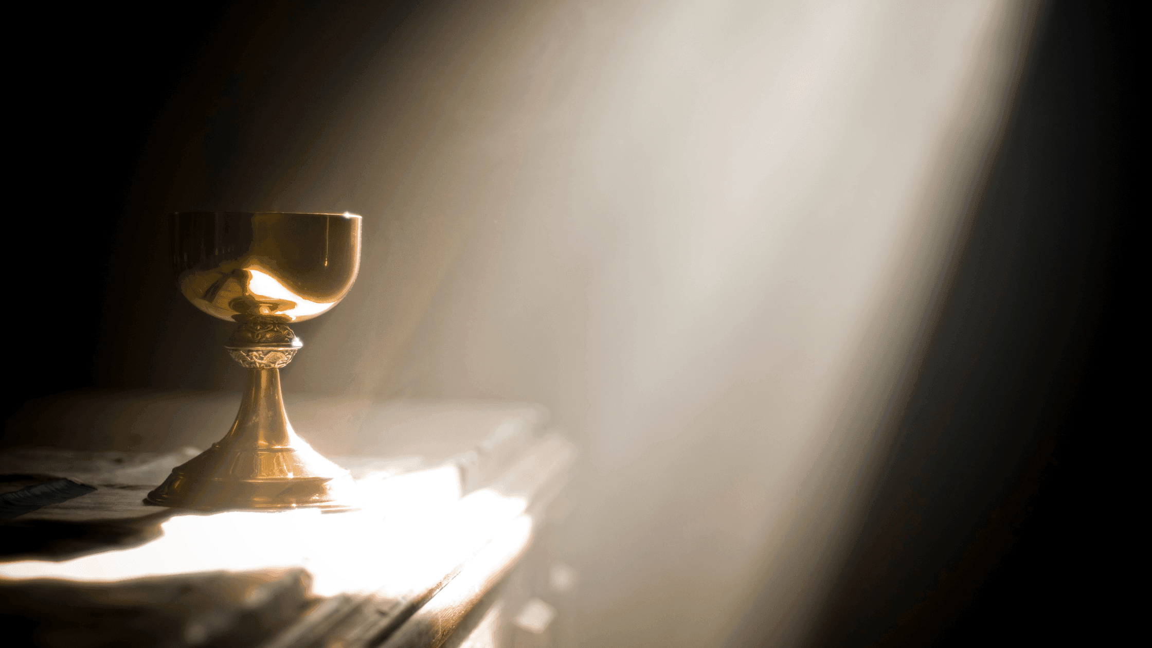 A golden chalice sits on a table, bathed in a beam of warm sunlight against a dark background, creating a serene and timeless atmosphere.