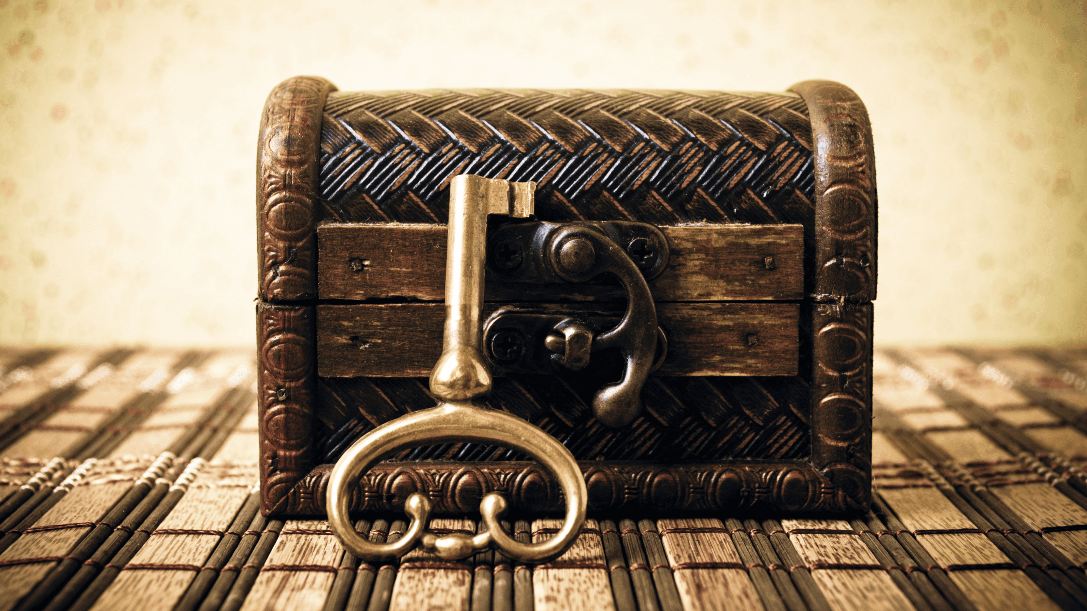 An ornate, vintage key rests against a small, decorative wooden chest with intricate patterns. The chest sits on a woven mat against a softly lit, textured background.