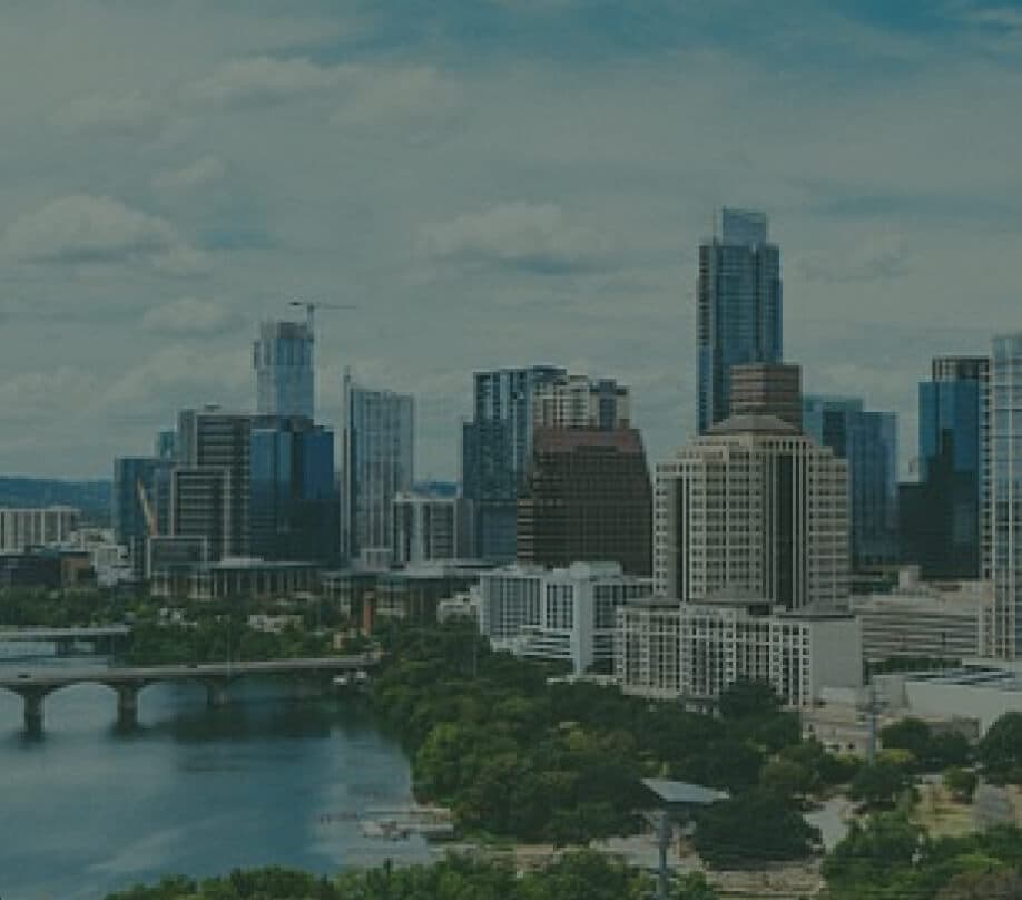 A scenic view of a city skyline featuring tall skyscrapers and modern buildings under a partly cloudy sky. In the foreground, there is a river with a bridge and lush green trees lining the waterfront.