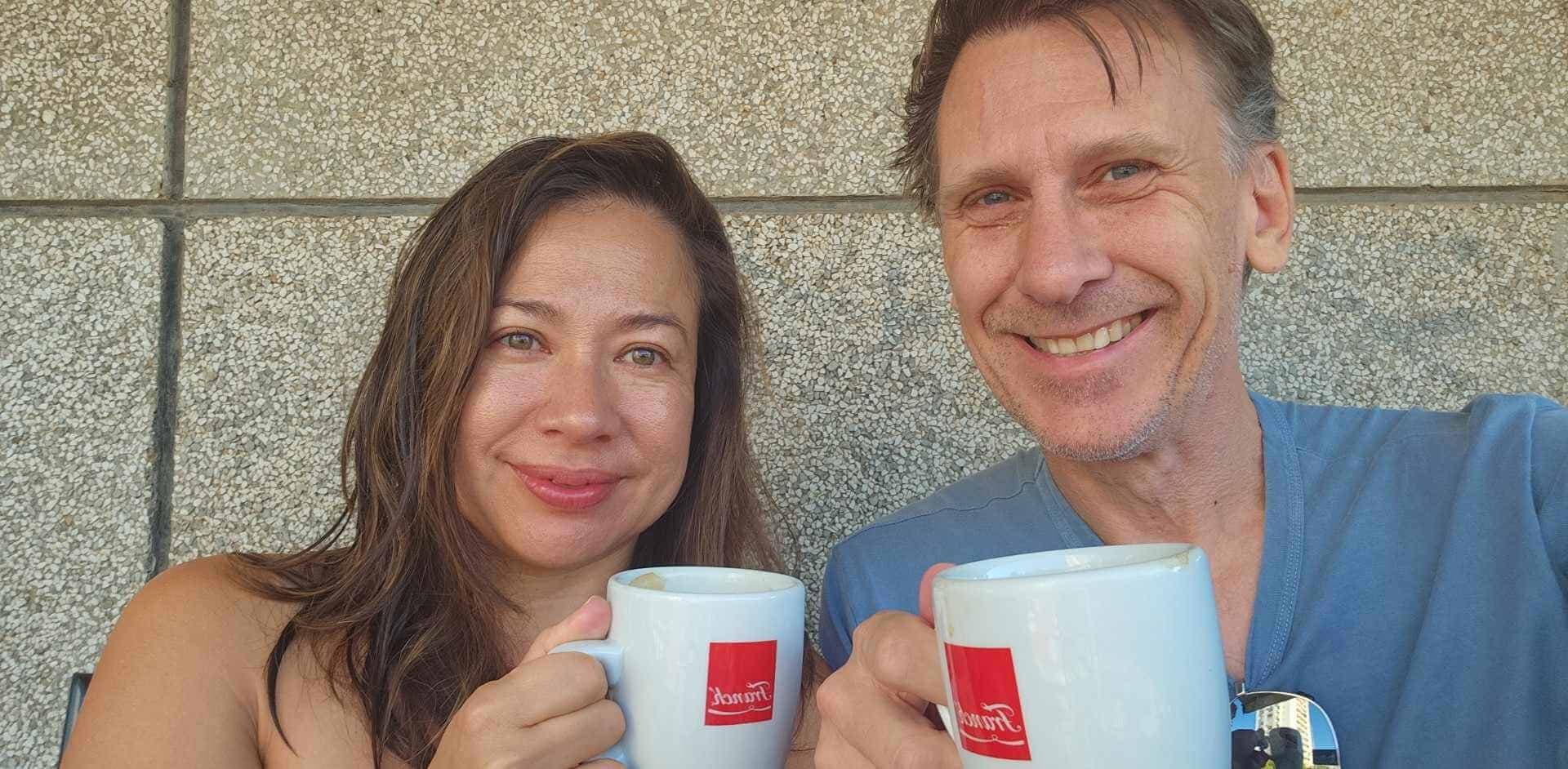 A man and woman smiling while holding white mugs with red logos in front of a textured wall. The man is wearing a blue shirt, and both appear to be enjoying a drink together.