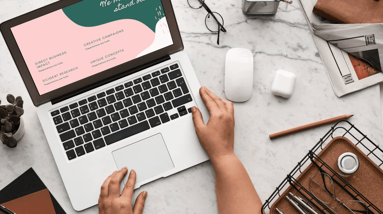 Person using a laptop with a pink presentation slide on the screen. The desk has glasses, a plant, earphones, a pen, and a notebook.