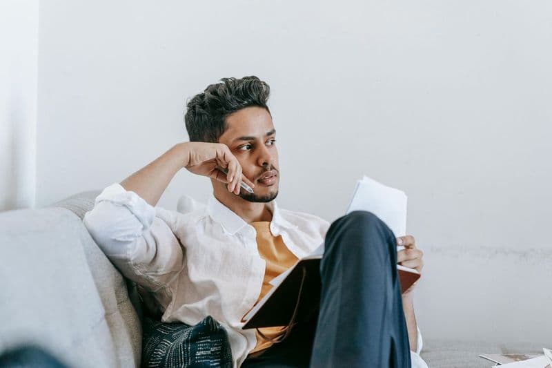 A man sitting on a couch, holding papers and a notebook, looks thoughtful with his hand resting on his face. He is wearing a white shirt and appears to be lost in contemplation.