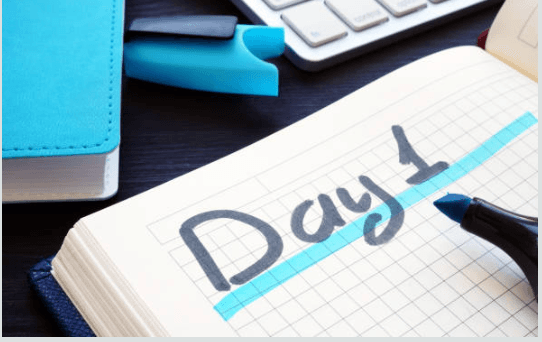 An open notebook shows Day 1 written in large letters with a blue marker. A keyboard, highlighters, and a closed blue notebook are nearby on a dark desk.