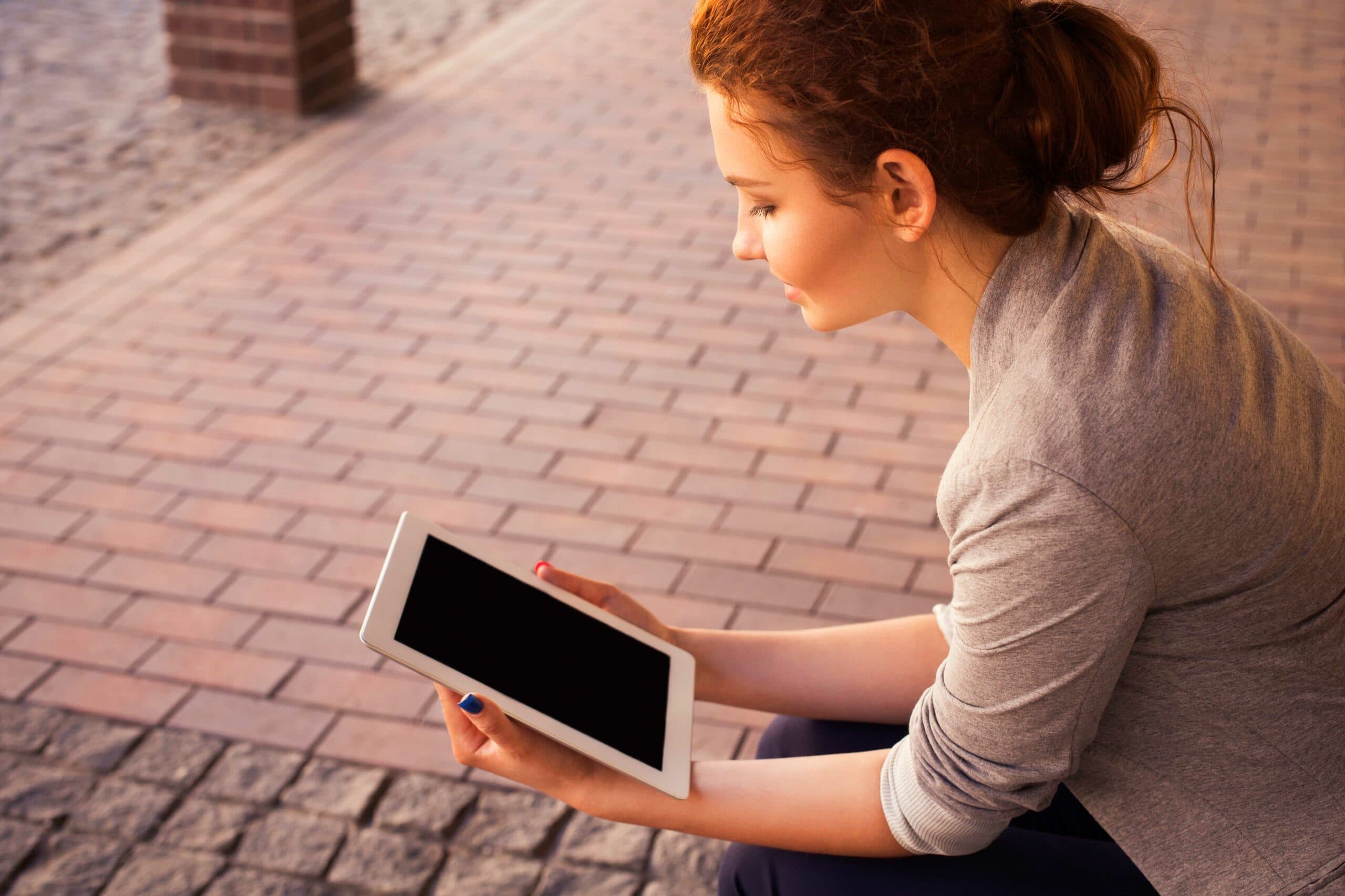 A person with brown hair tied back is sitting outdoors on a brick pavement, holding a tablet. They are wearing a gray sweater and have a blue nail polish on one hand. The screen of the tablet is visible but turned off.