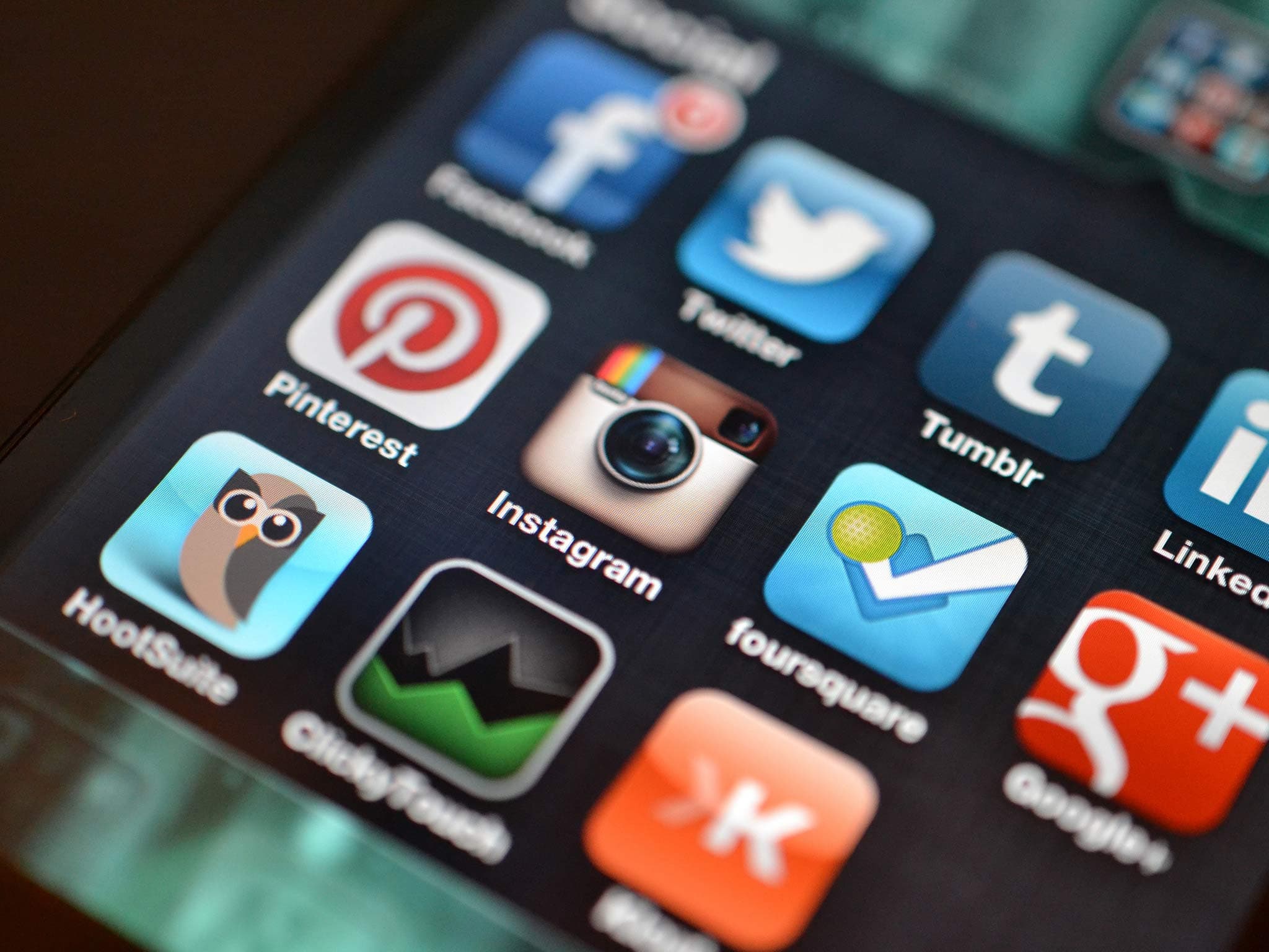 Close-up of a smartphone screen displaying various social media app icons, including Facebook, Twitter, Pinterest, Instagram, Tumblr, Hootsuite, and Foursquare, arranged in a grid layout. The background is darkened.