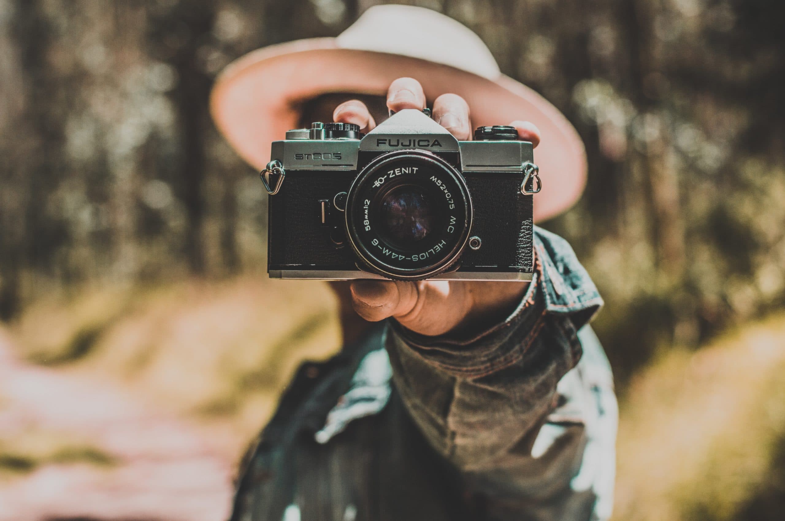 A person wearing a hat holds a vintage Fujica camera towards the viewer, with a blurred natural background. The focus is on the camera, emphasizing its lens and details.