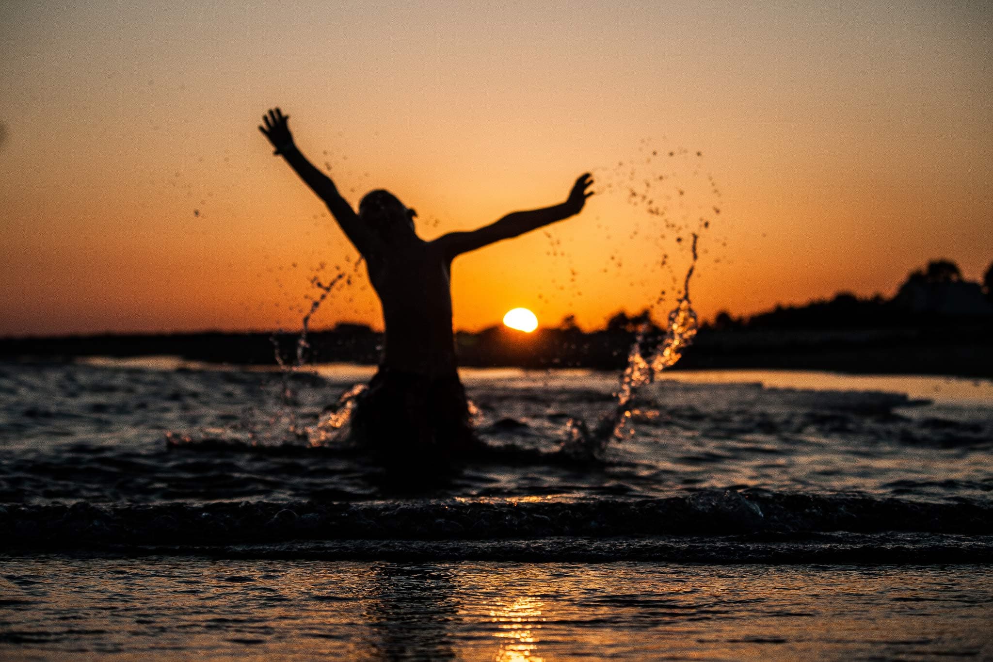 A person silhouetted against a sunset splashes in ocean waves with arms outstretched, creating water arcs. The horizon is visible in the background, and the scene exudes a joyful, carefree atmosphere.