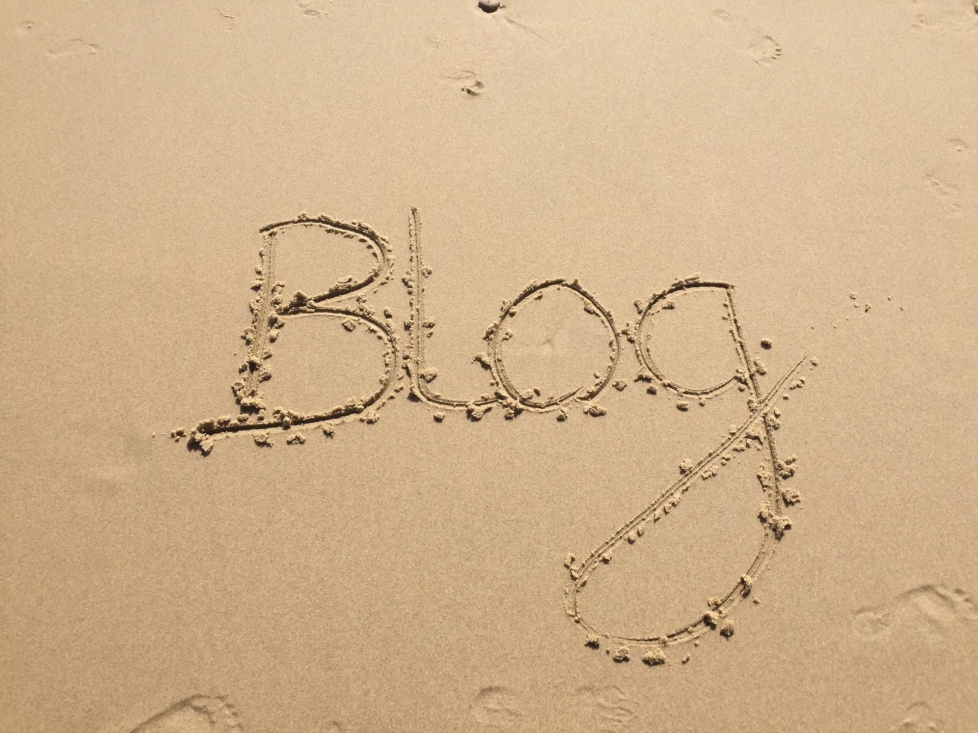 The word Blog is written in cursive in the sand on a beach, with footprints visible nearby.