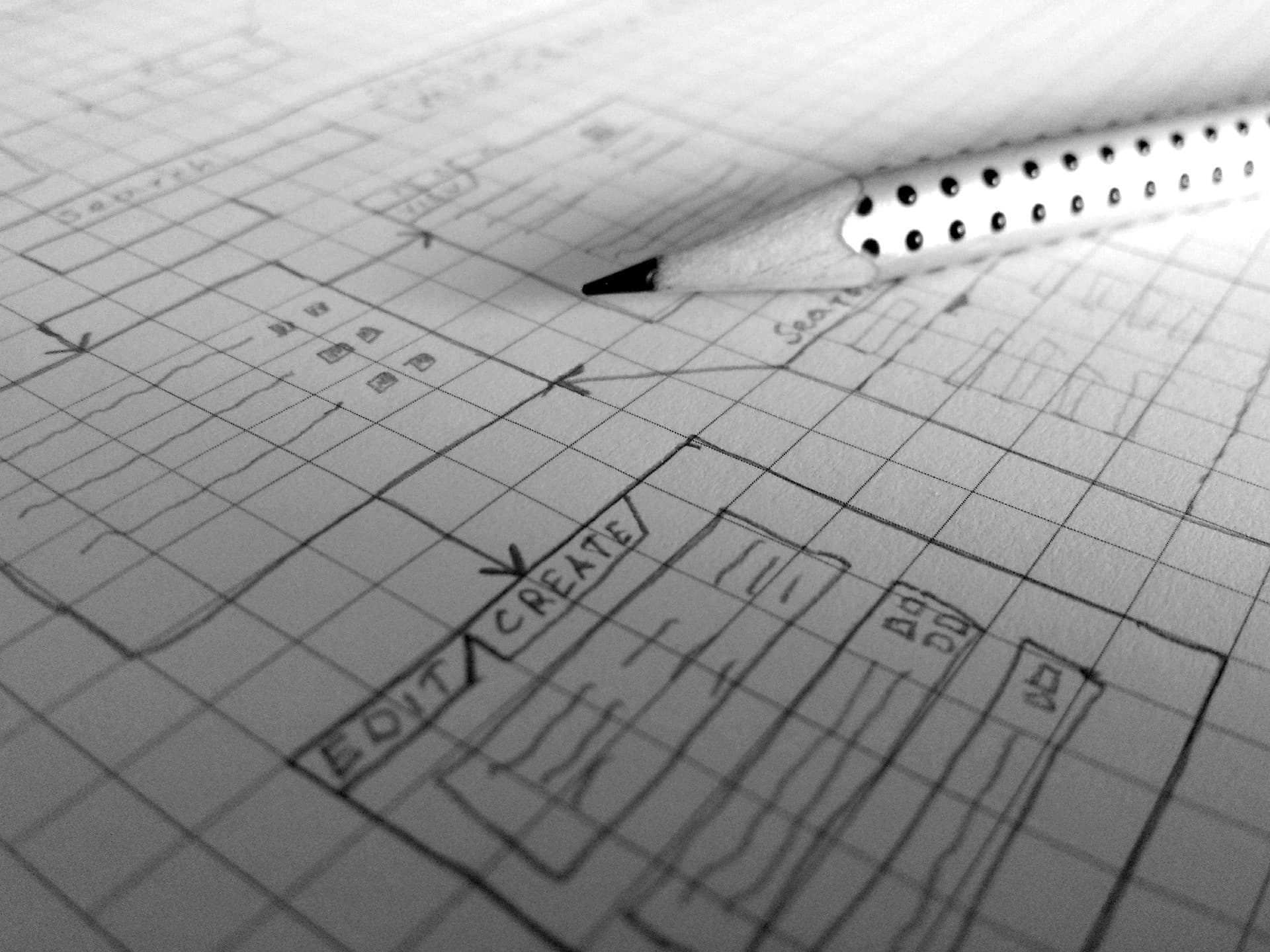 A black and white photo of a pencil resting on graph paper with a sketched design. The paper features boxes and words like EDIT and CREATE, indicating a layout or blueprint.