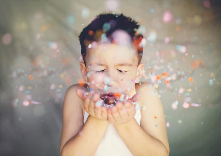 A young child with closed eyes blows a handful of colorful confetti, creating a vibrant burst of colors in the air. The child is wearing a sleeveless shirt and stands against a soft-focus background.