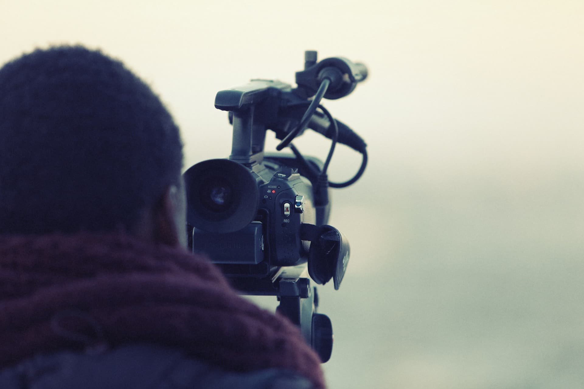 A person stands behind a professional video camera, which is focused on a distant scene. The person is wearing a scarf and the atmosphere appears calm and misty, suggesting early morning or late evening.