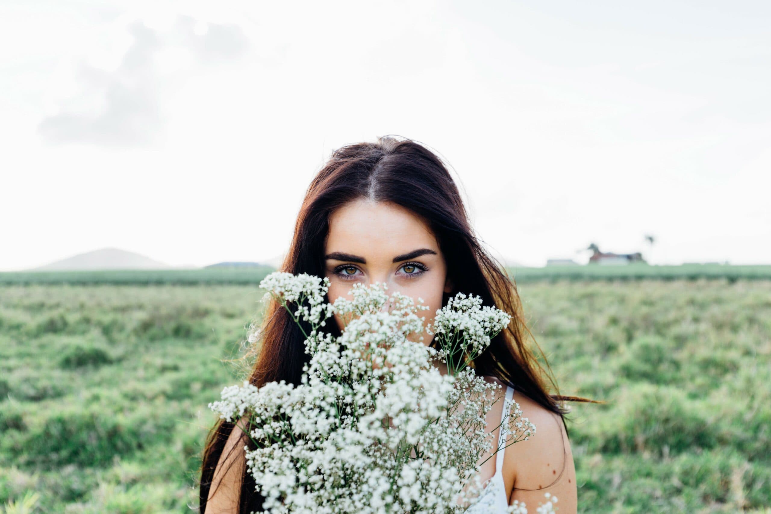 A woman with long dark hair stands in a grassy field, partially obscured by white flowers she holds. Her piercing eyes gaze directly at the camera, with an expansive, blurred landscape in the background.