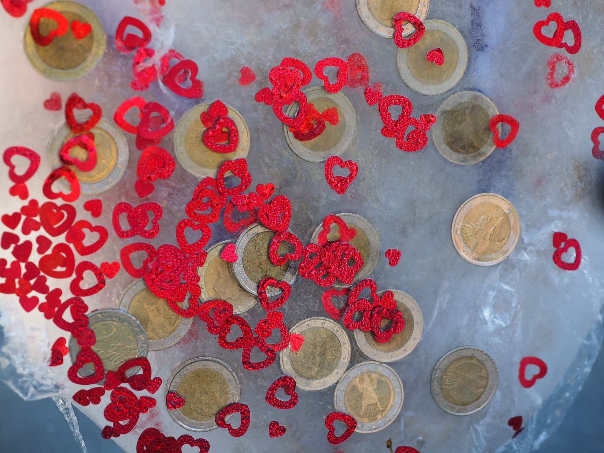 Scattered Euro coins on a transparent surface surrounded by small red heart-shaped confetti. The background is blurred, highlighting the mix of shiny metallic coins and vibrant confetti.