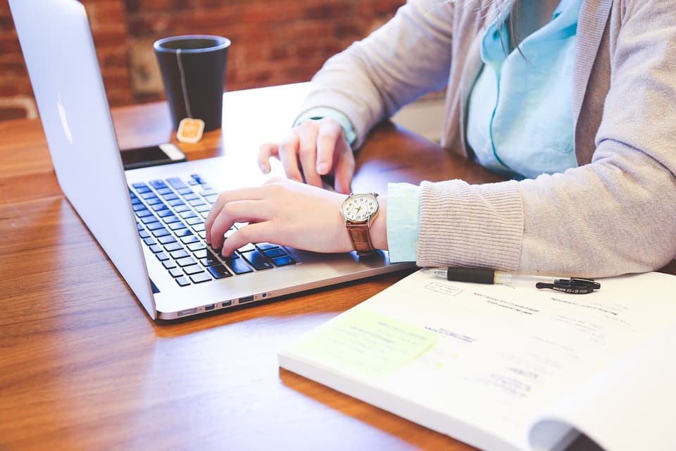 Person typing on a laptop at a wooden table, with a notebook, pen, and a takeaway coffee cup nearby. Theyre wearing a light sweater and a watch. The brick wall in the background gives a cozy atmosphere.