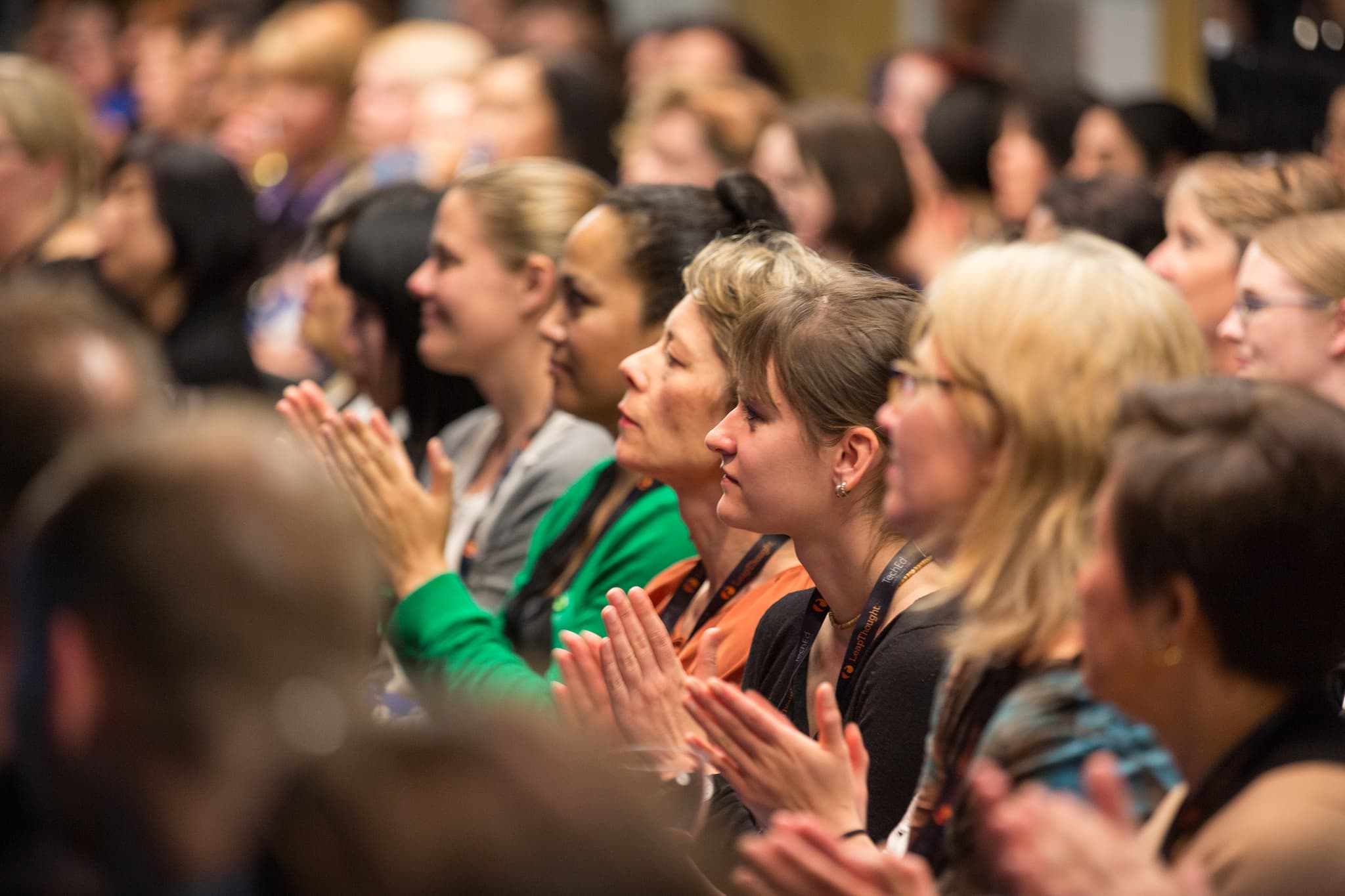 An audience of people sits closely together, facing forward. Many are clapping, and their expressions reflect engagement. The crowd is composed of adults of various ages, with a mix of clothing styles and colors.