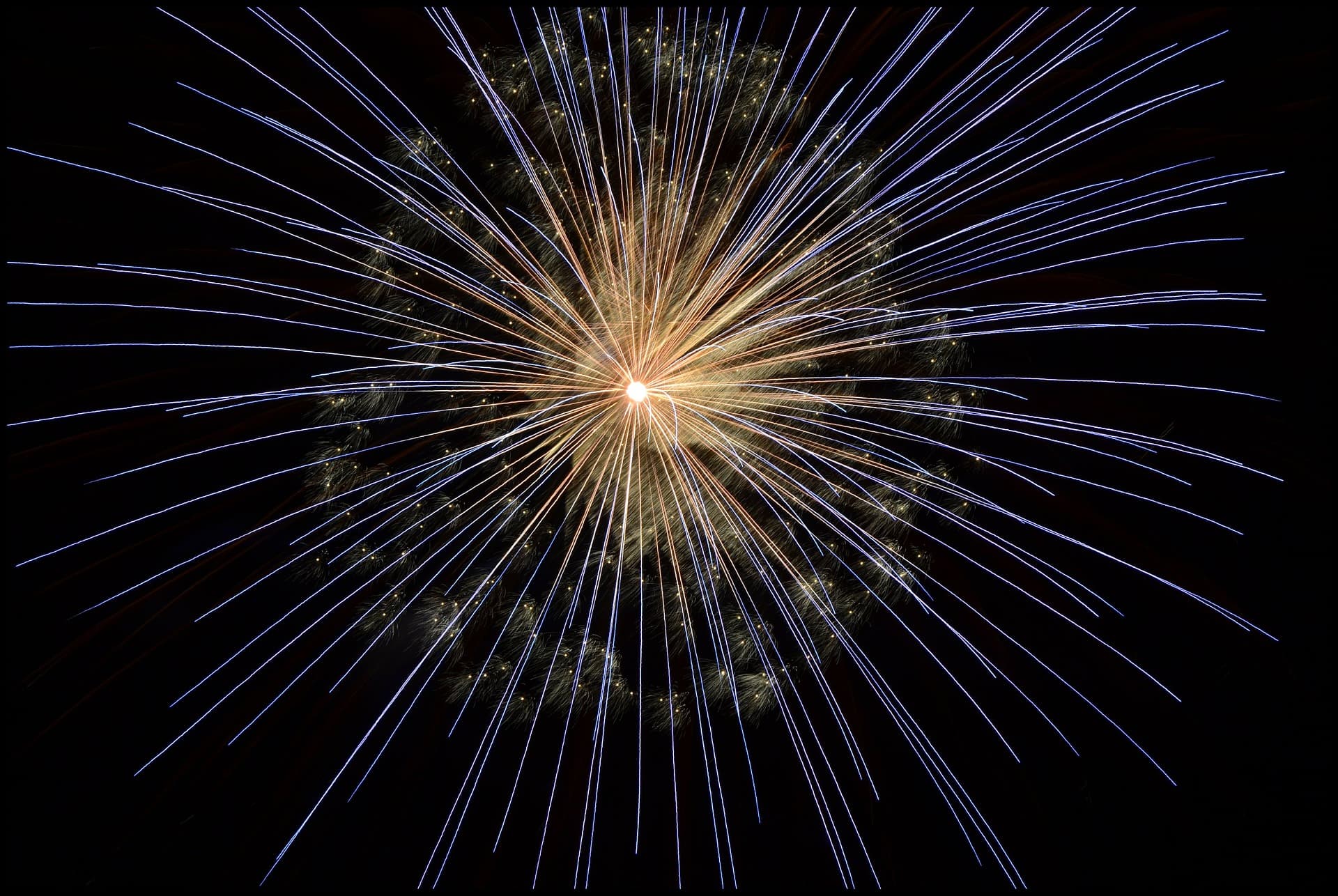 A circular burst of fireworks radiates from a bright central point against a dark night sky. The display features long, thin streaks of white and blue light, creating a star-like pattern.