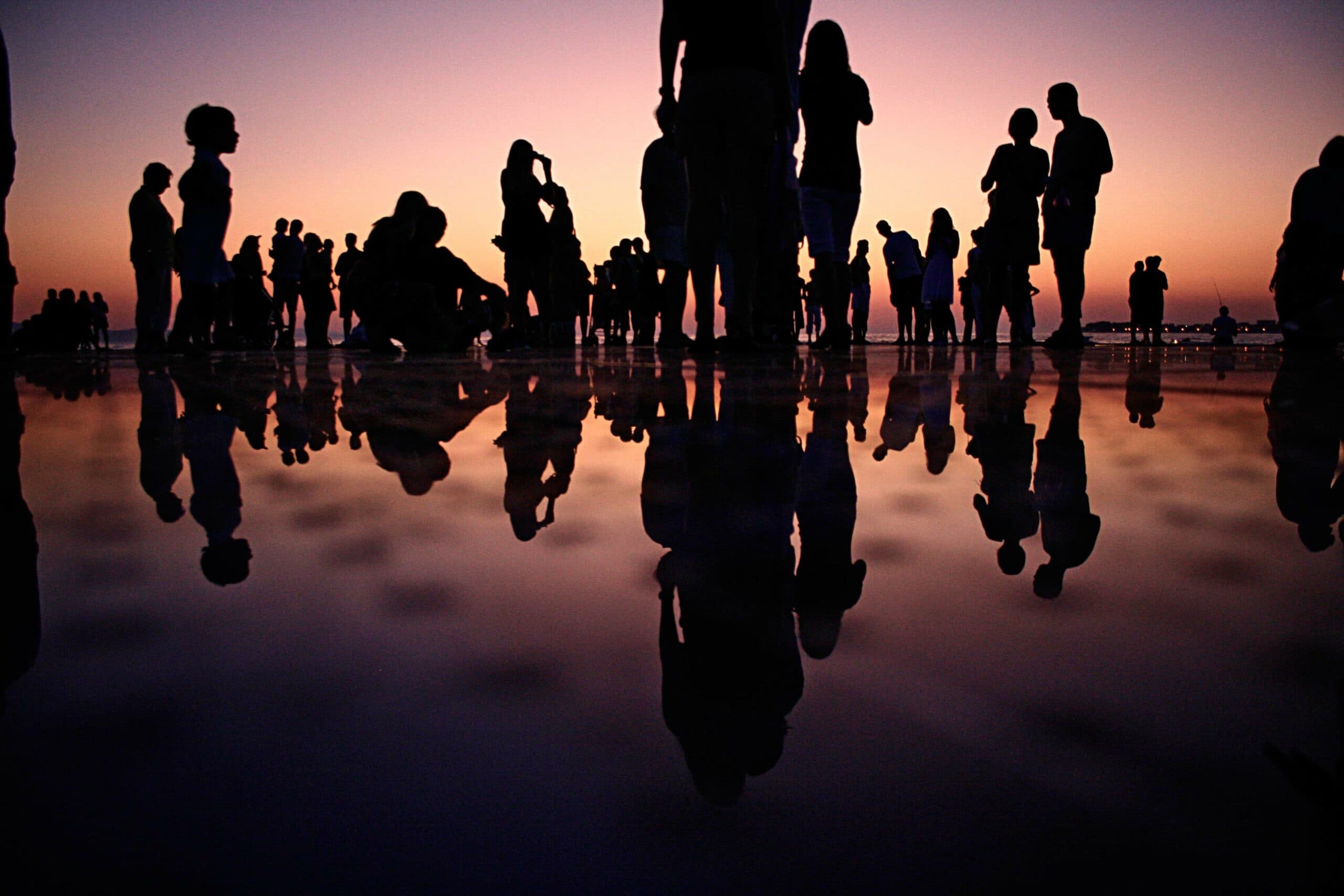 Silhouettes of people gathered on a reflective surface at sunset. The sky is a gradient of warm colors, and the group stands in various poses, creating mirrored reflections on the wet ground.