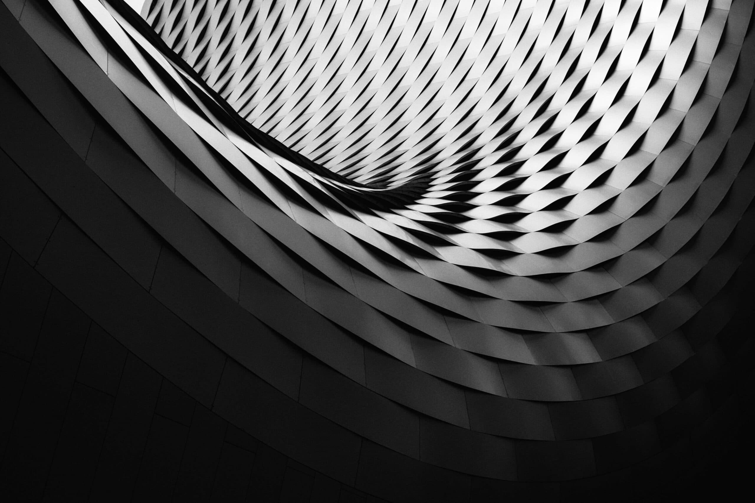 A dramatic black and white image of a building with a swirling, wavy pattern. The structure features a repeating grid of diamond-shaped panels, creating a dynamic, flowing architectural design that draws the eye upward toward the light.