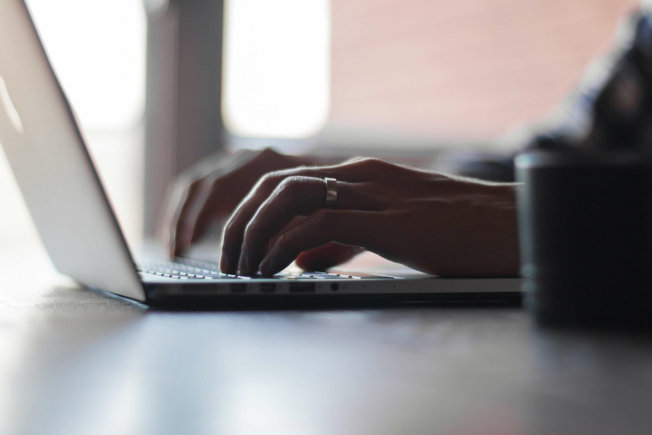 Close-up of a person typing on a laptop keyboard. The persons hands are visible, with a ring on the left hand. The scene is softly lit, creating a calm and focused atmosphere. A blurred background emphasizes the hands and keyboard.