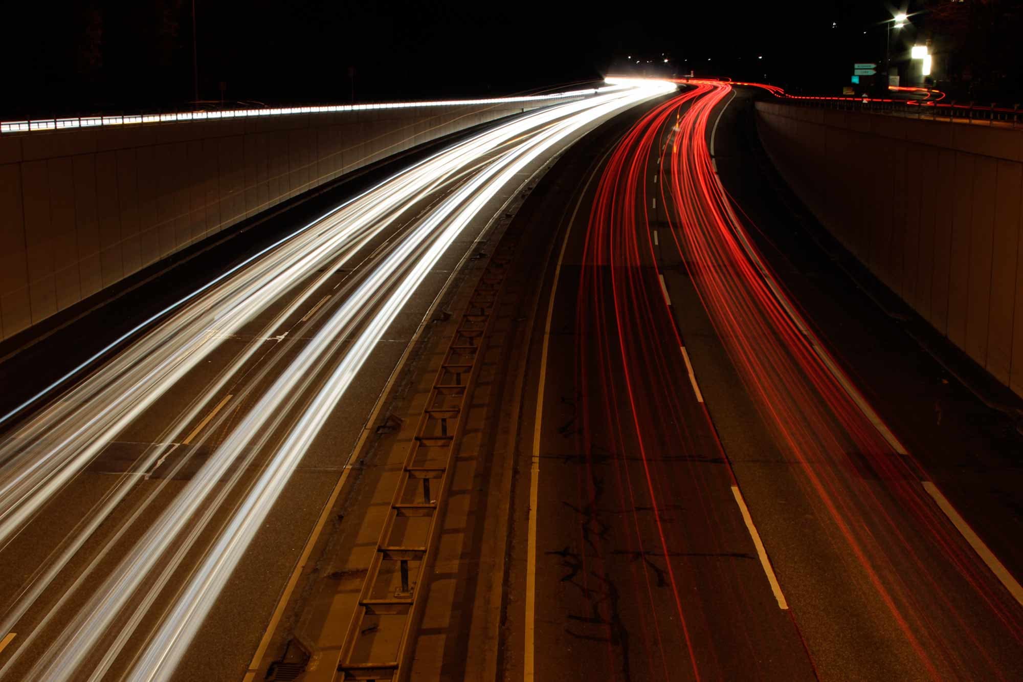 Long exposure photo of a highway at night, showing white and red light trails from vehicle headlights and taillights. The lanes are separated by a barrier, with the red lights on the left and white lights on the right.